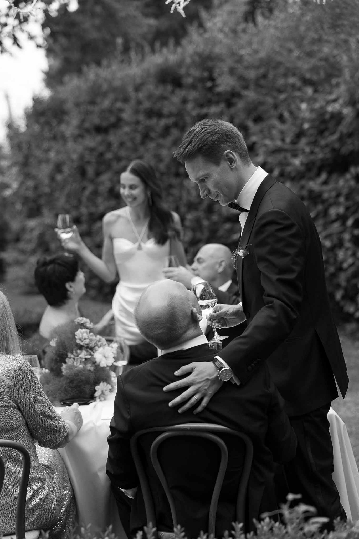 Black and white candid of groom clinking glasses with guest while bride raises glass at garden table