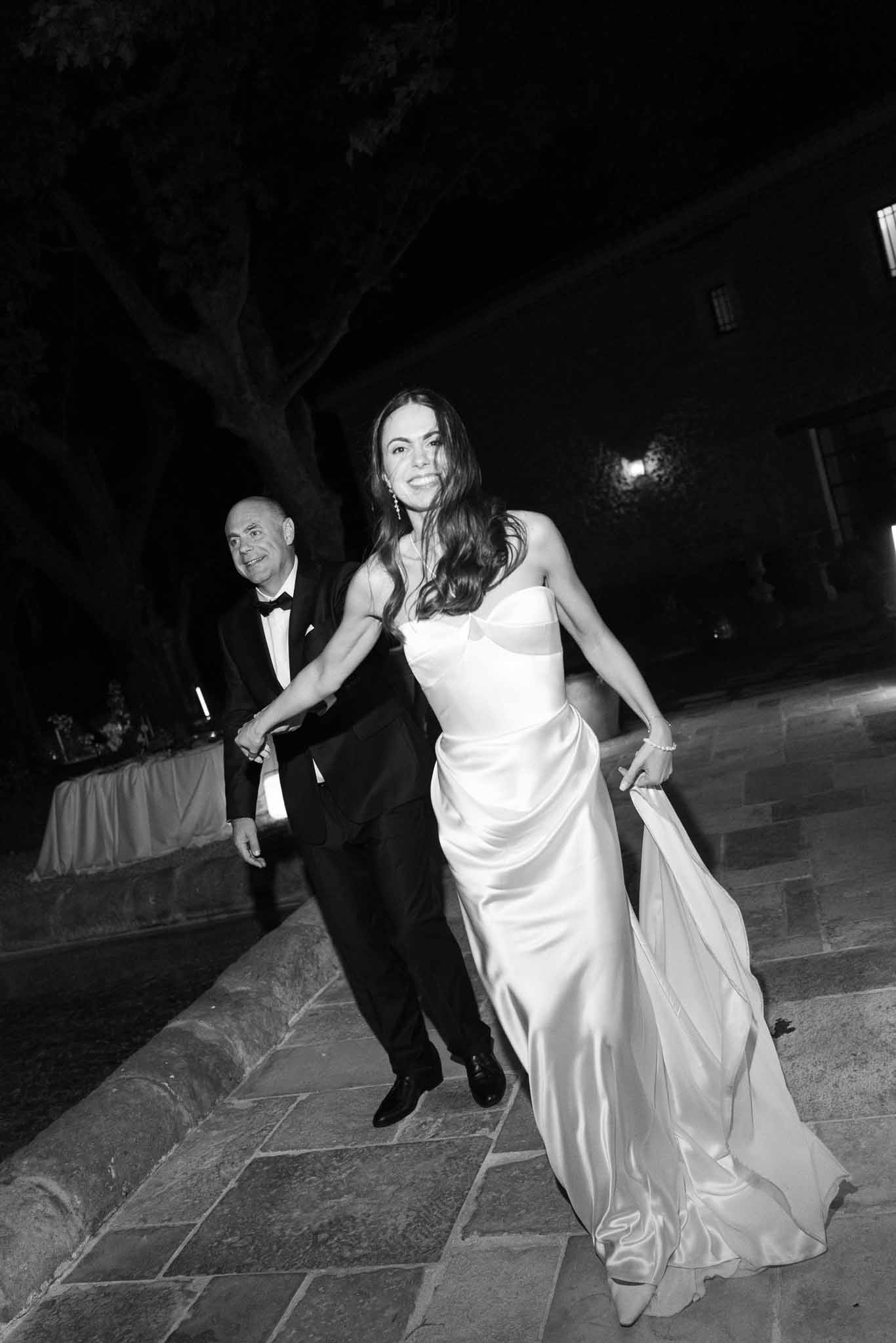 Black-and-white nighttime photo of bride in strapless satin gown walking with her father along a stone path