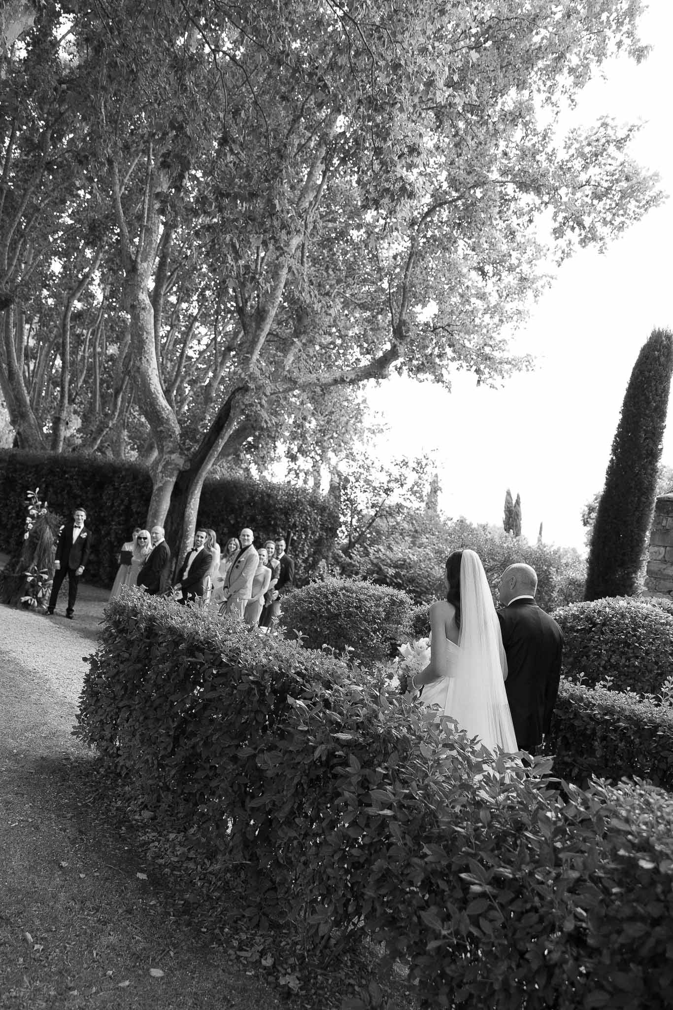 Bride escorted down gravel path through clipped box hedges toward waiting groom and guests in B&W