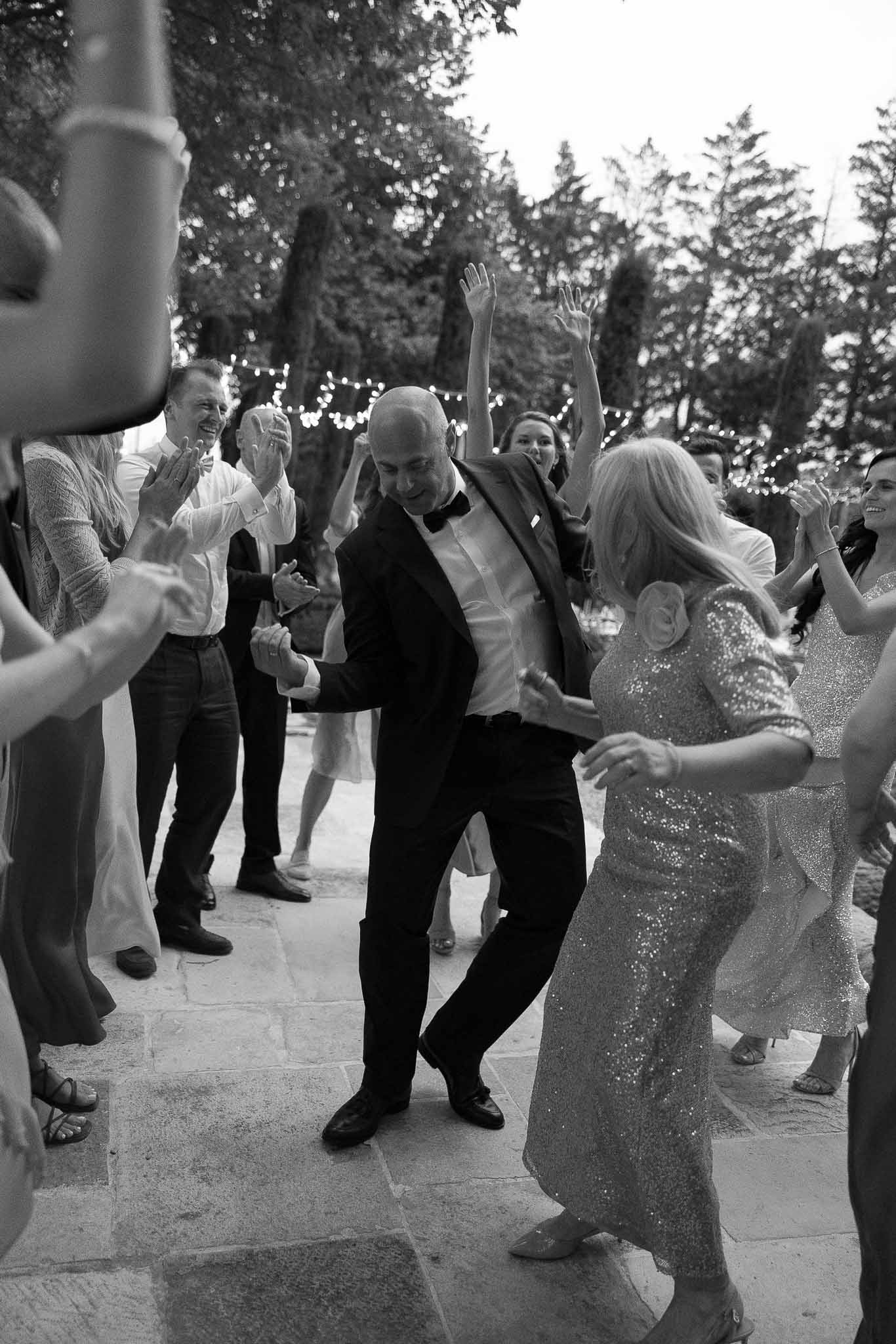 Black and white of guests dancing energetically on stone terrace with fairy lights at evening reception party
