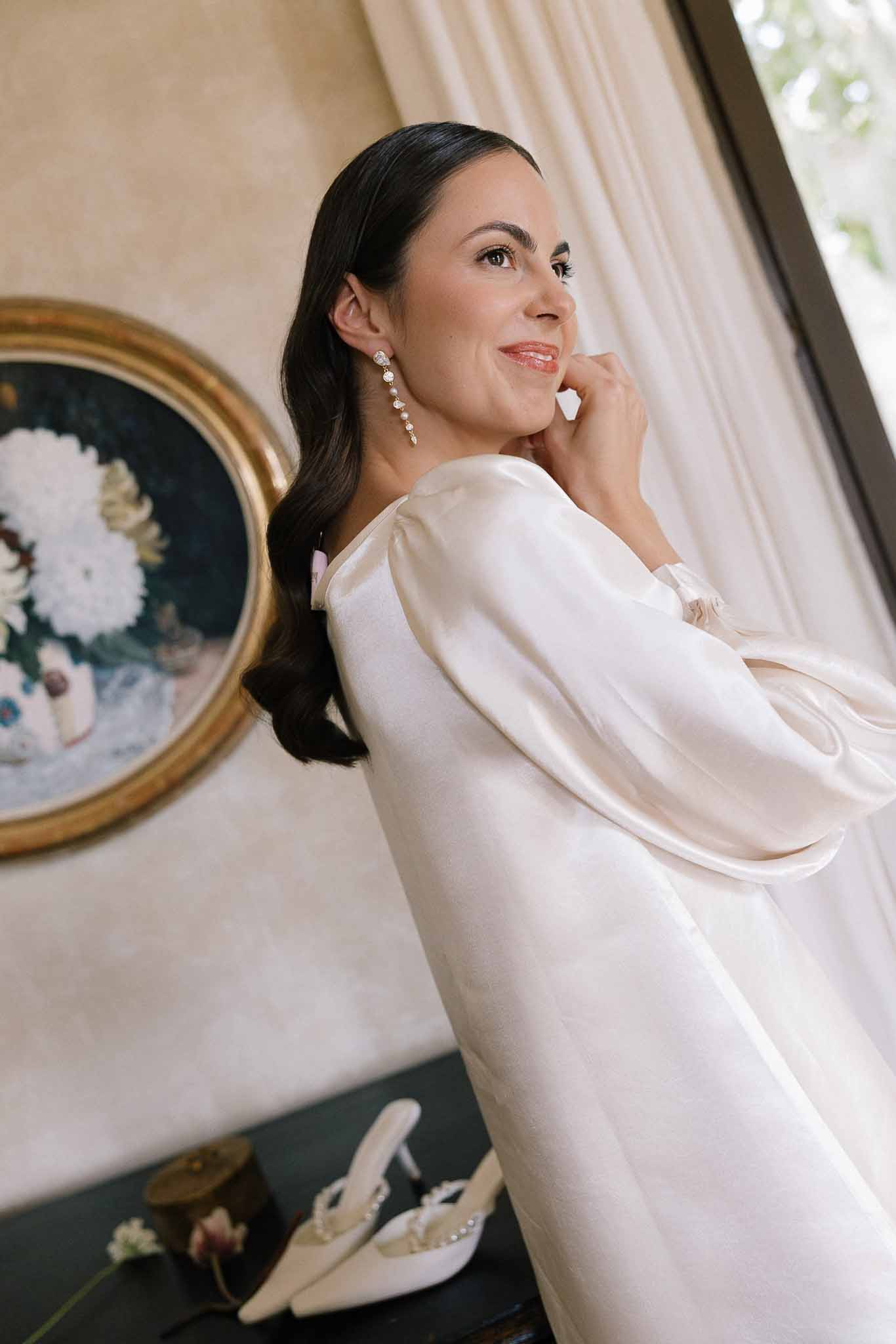 Bride in cream satin robe with crystal earrings and side-wave hair smiling beside gold-framed painting