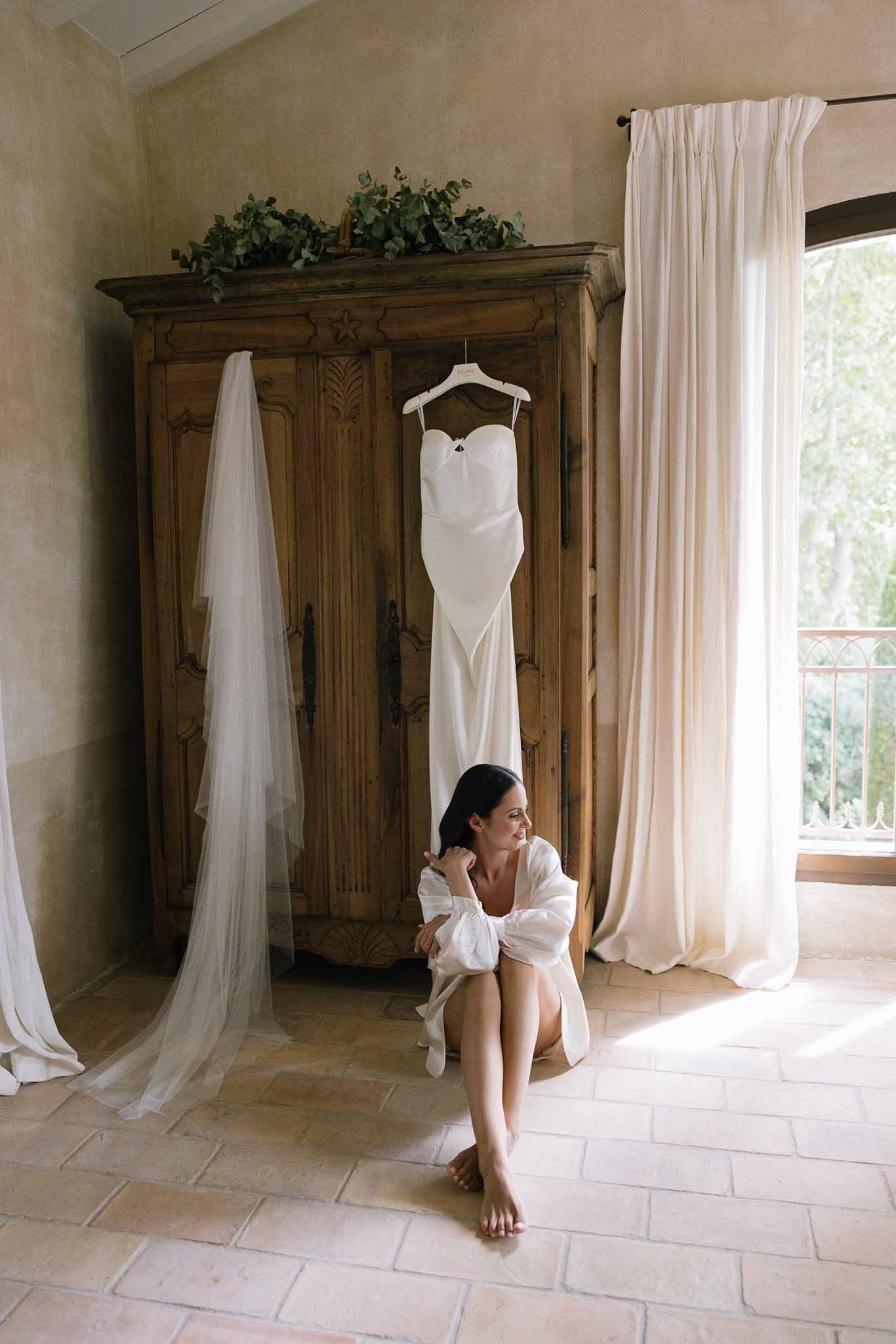 Bride in white robe seated on floor below ivory gown and cathedral veil hanging from carved armoire