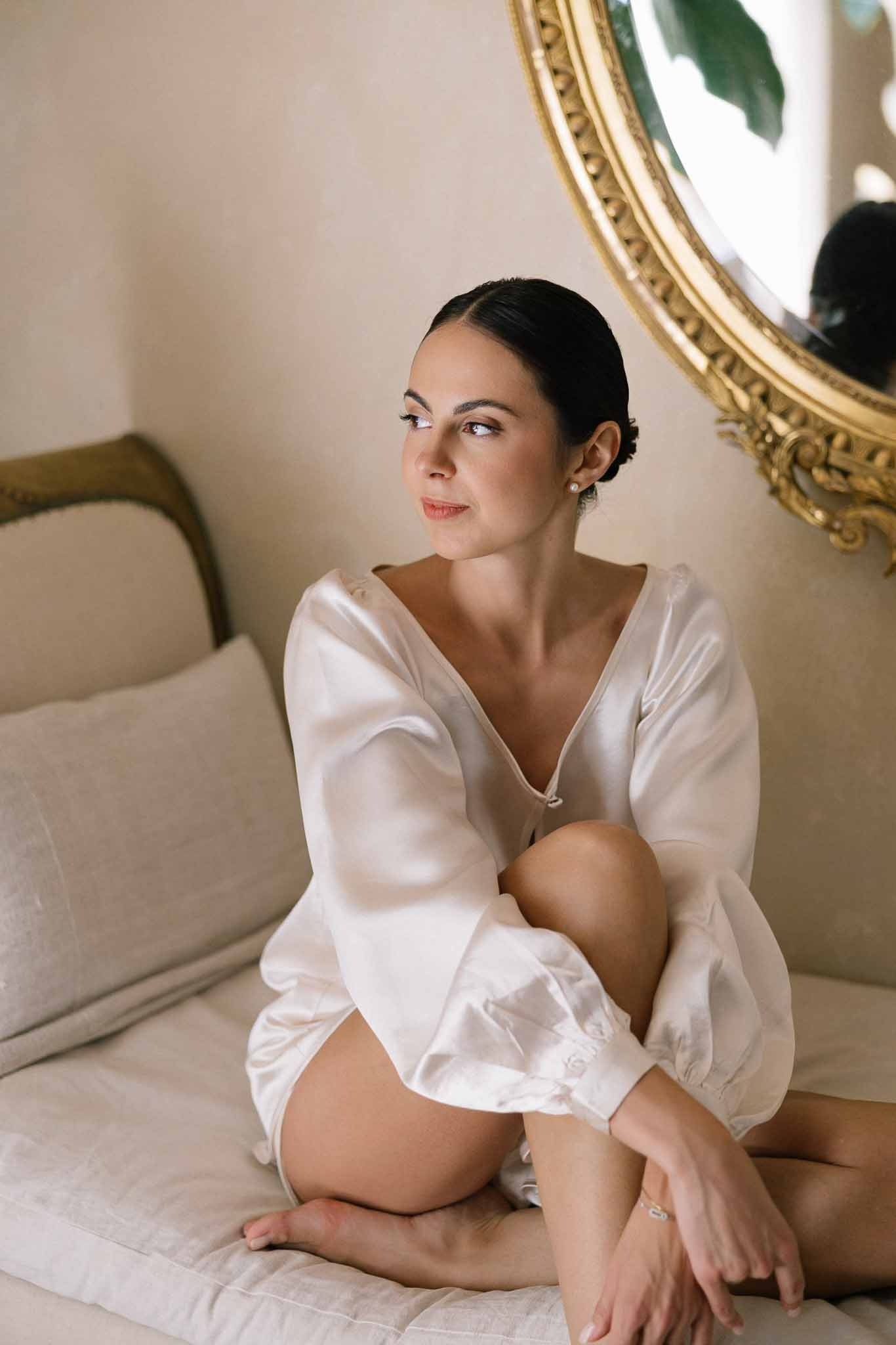 Bride in ivory satin robe with sleek low bun seated on daybed before ornate gold-framed oval mirror
