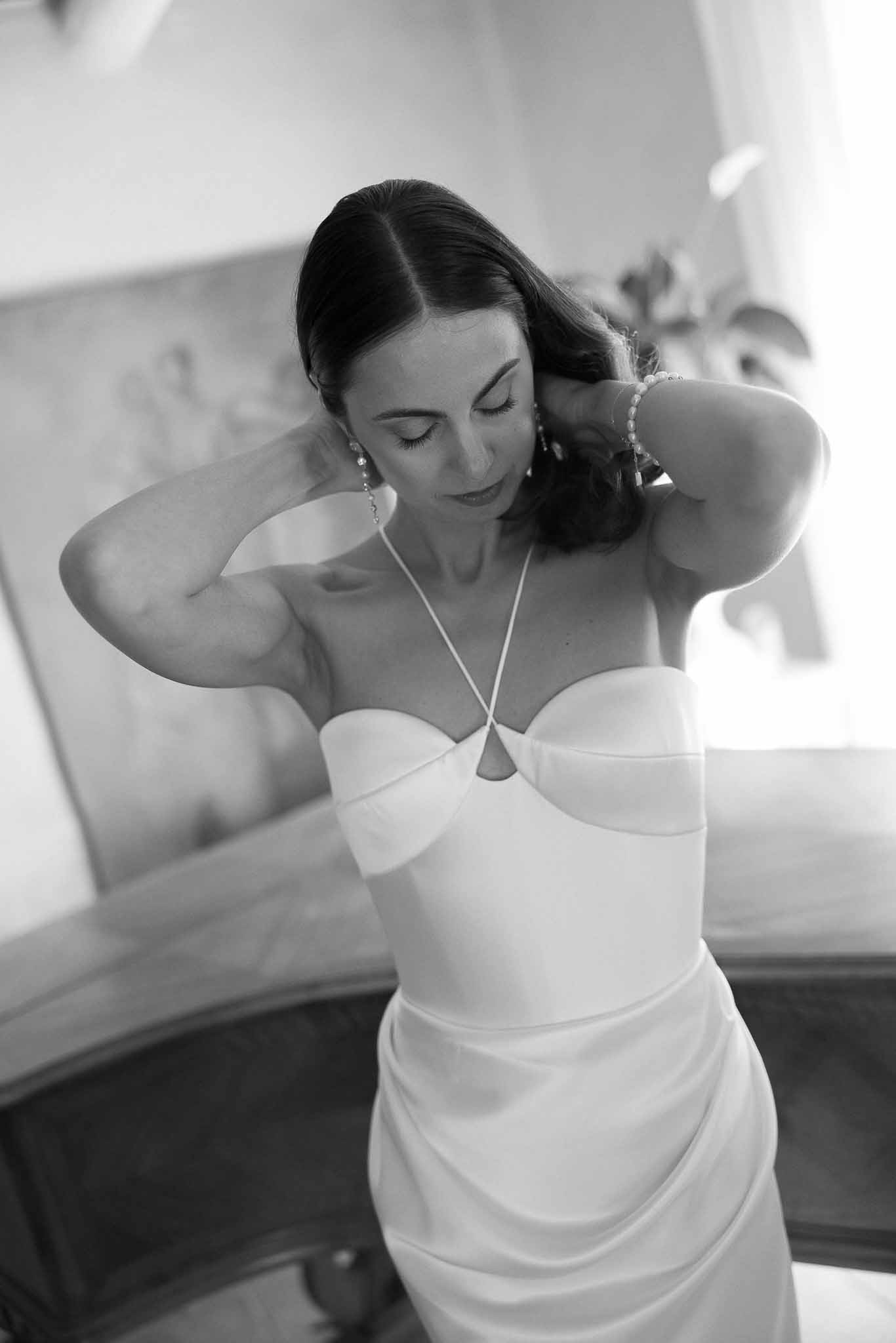 Black-and-white portrait of bride fastening necklace while wearing halter-style wedding dress with sweetheart bodice