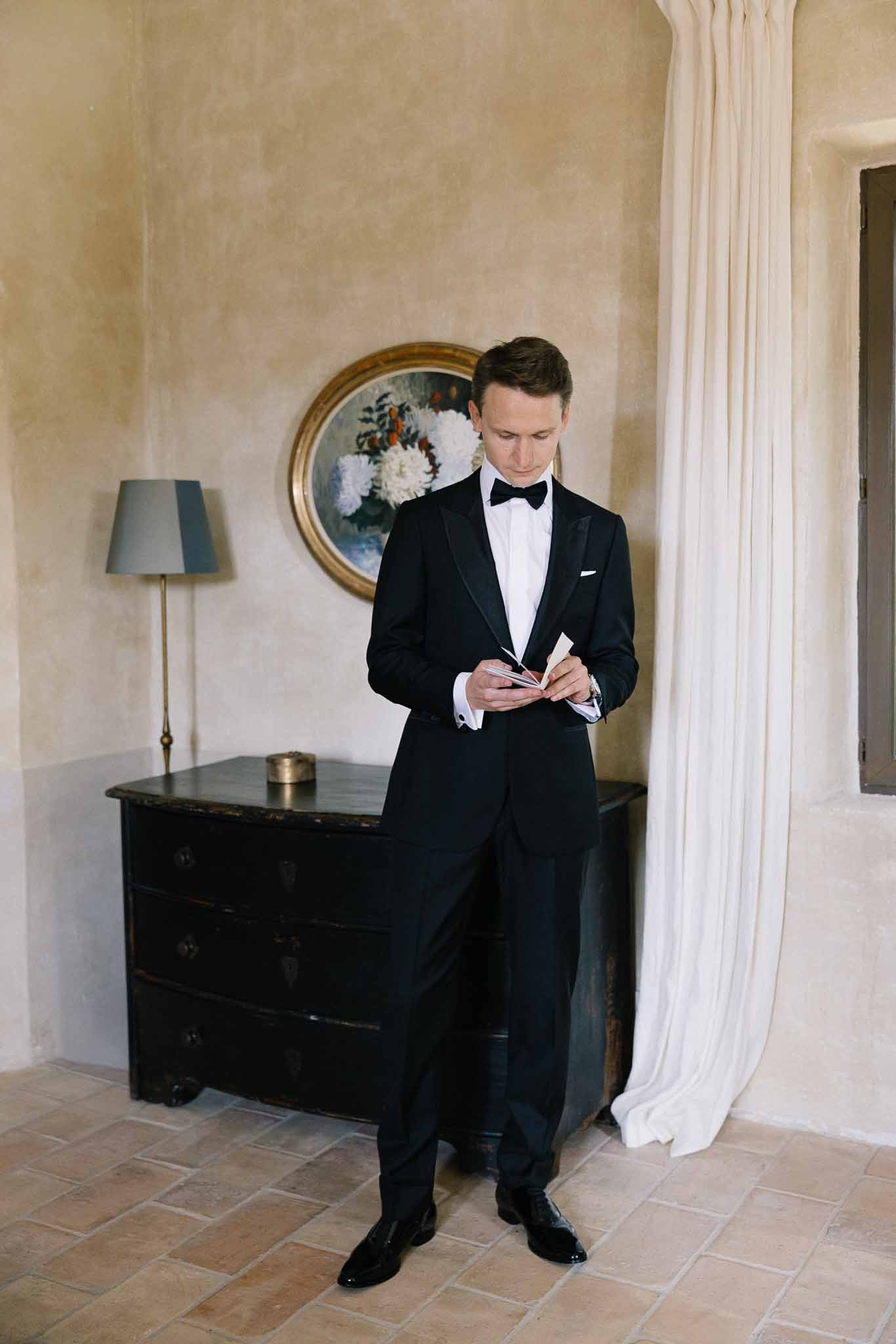 Groom in black tuxedo reading card in ochre-walled room with brass lamp and gold-framed painting