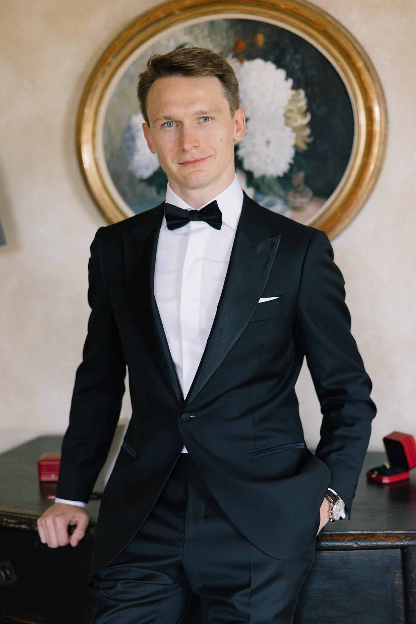 Groom in black tuxedo with bow tie leaning against dresser with Cartier ring boxes and oil painting