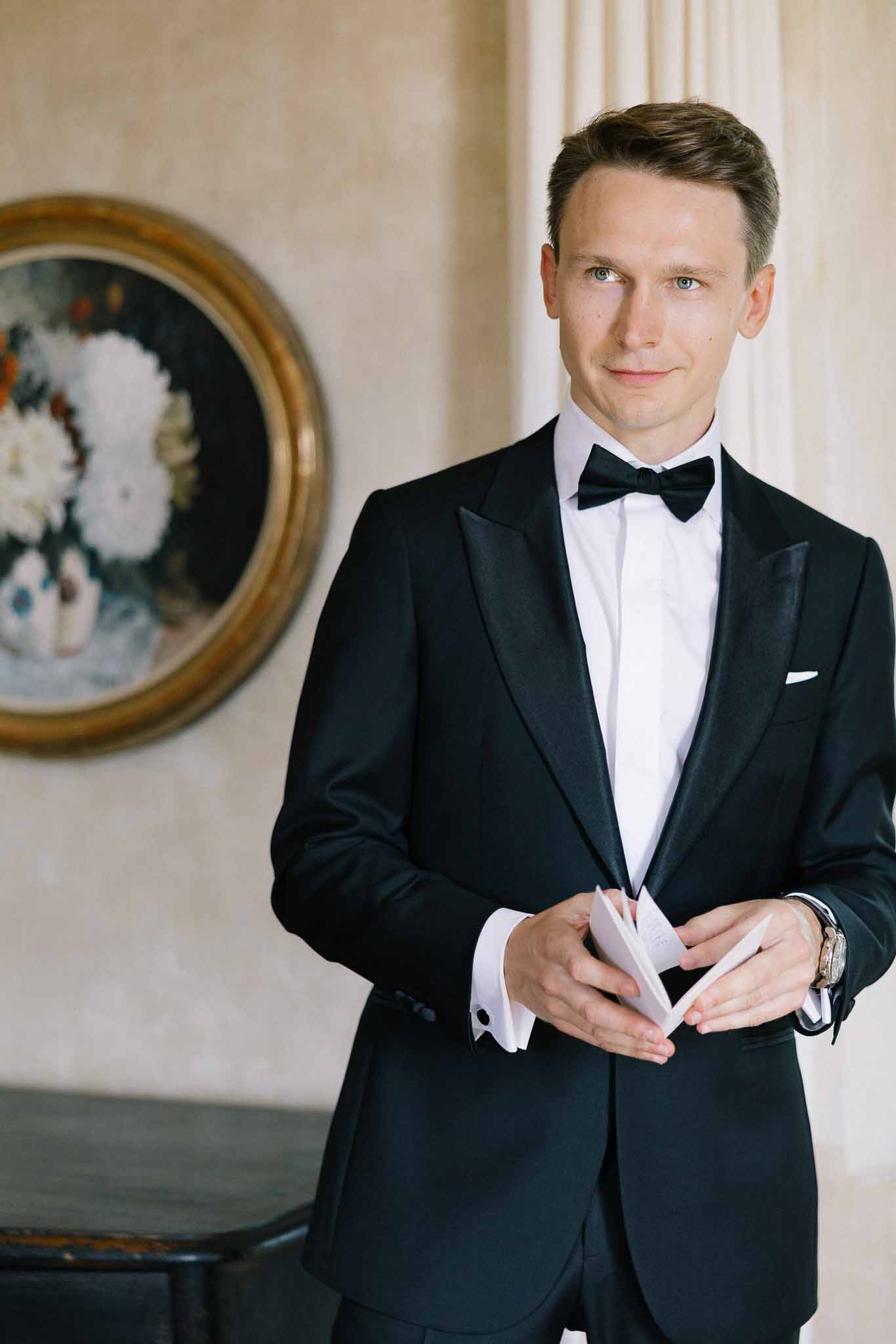 Groom in black tuxedo with bow tie holding a note in chateau room with gold-framed painting