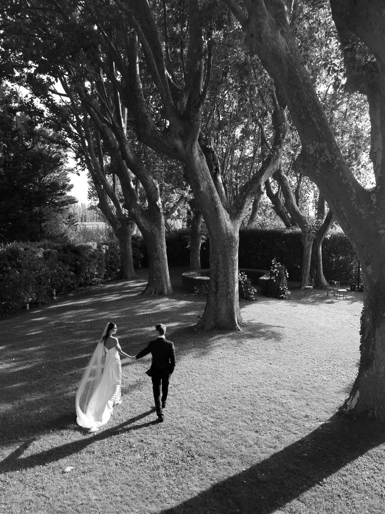Bride and groom walking hand-in-hand through tree-lined garden with cathedral veil trailing in black and white
