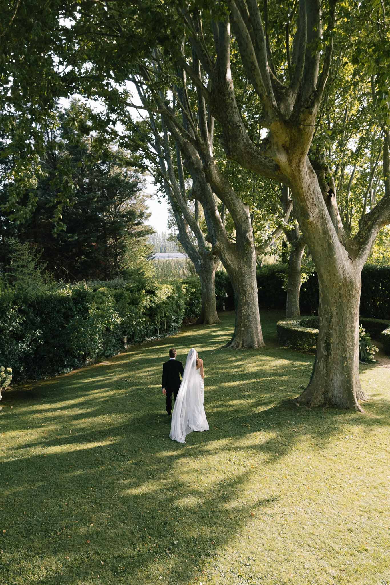 Elevated view of couple walking across formal garden with plane trees and topiary, cathedral veil trailing on grass