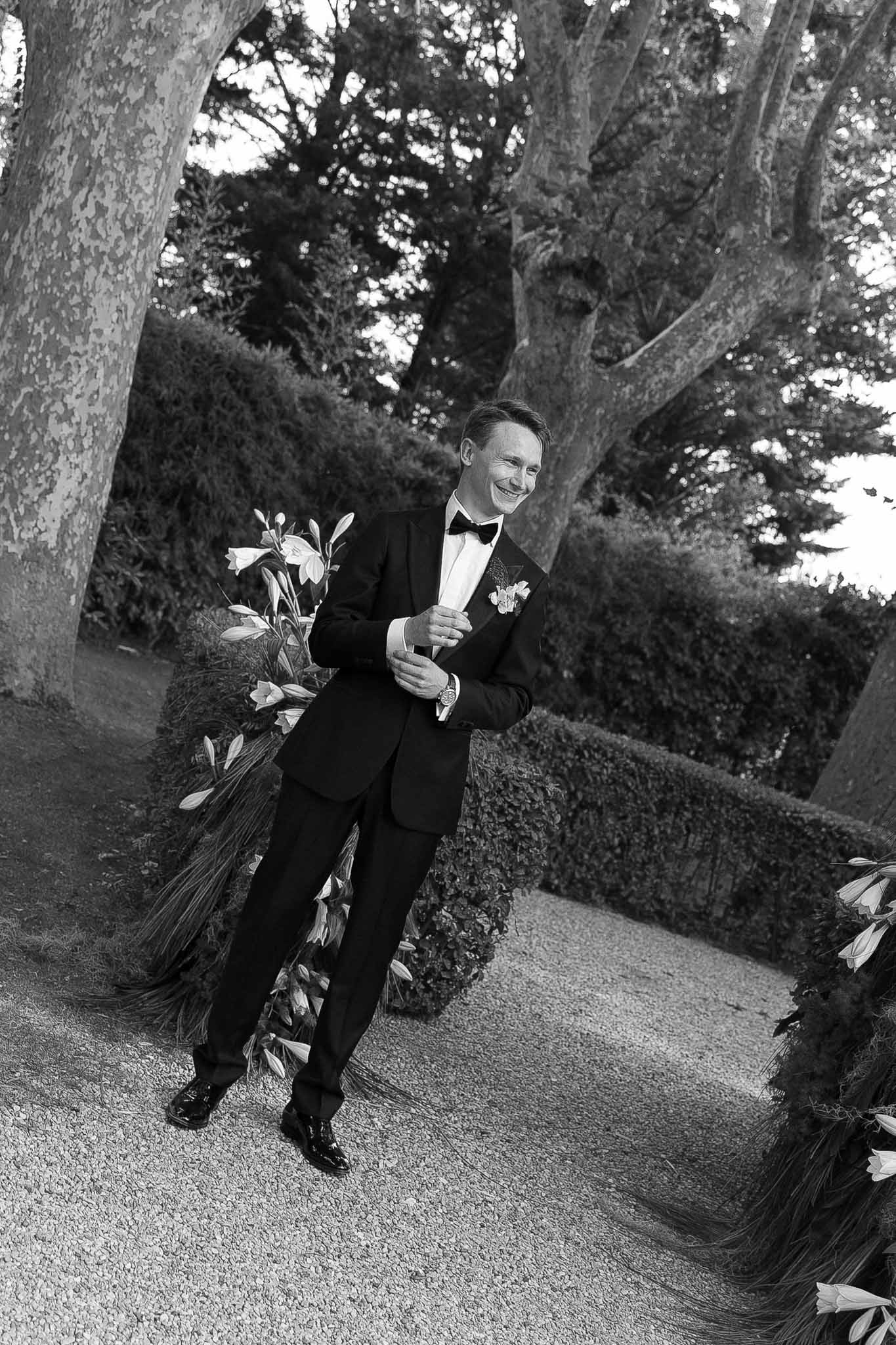 Black and white groom laughing on gravel path adjusting cuff with lily arrangements flanking ceremony aisle