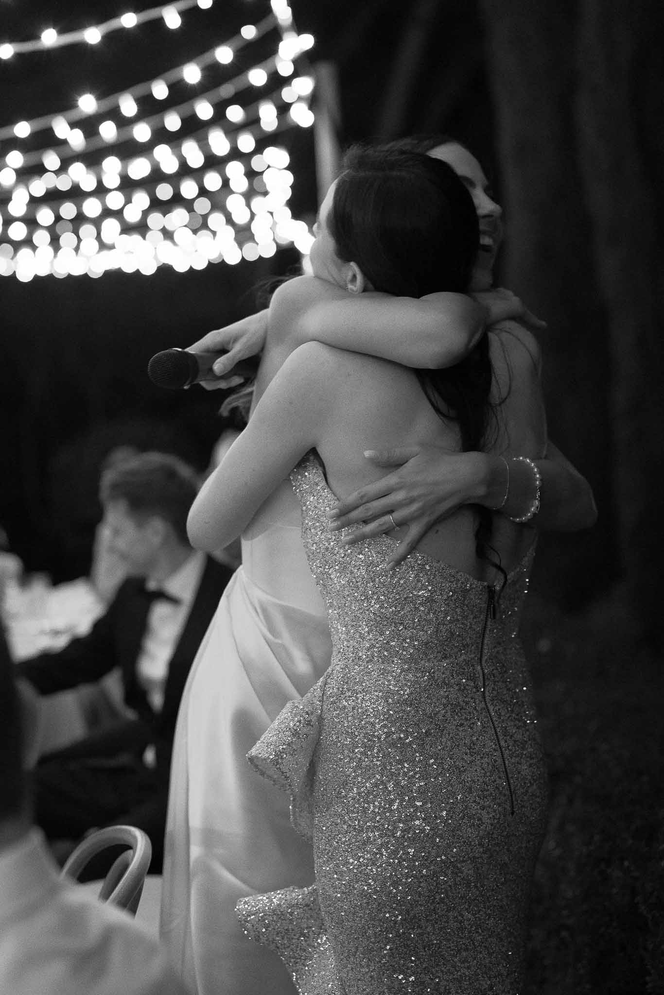Black and white bride embracing friend after speech with fairy light bokeh overhead