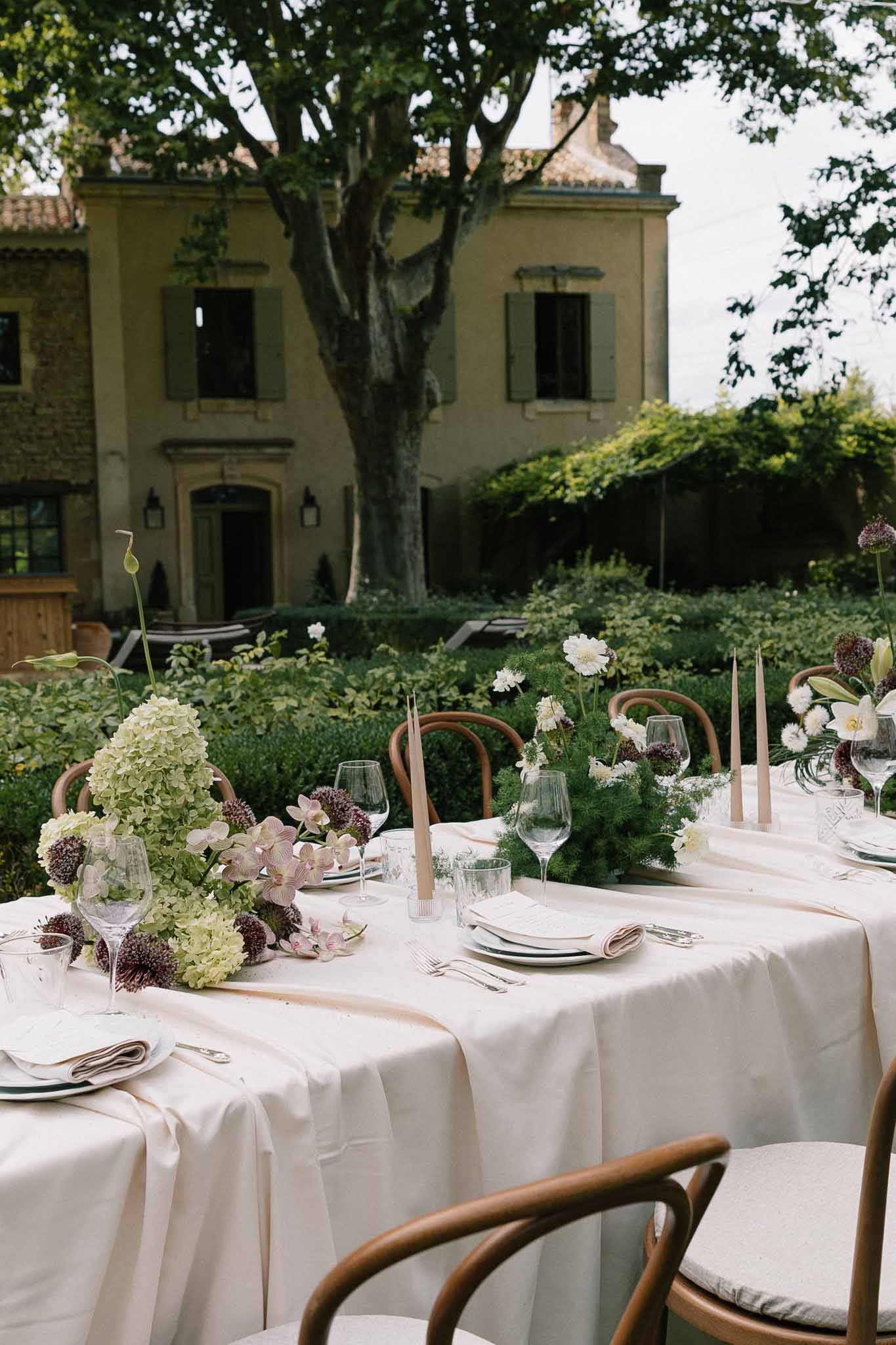 Reception table with allium orchid and dahlia runner and crossback chairs before yellow manor house