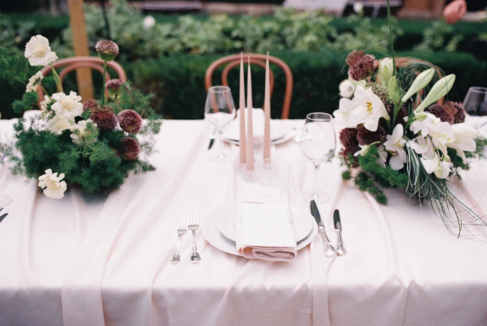 Outdoor reception place setting with blush linen, white plates, menu card, taper candles and white and burgundy florals