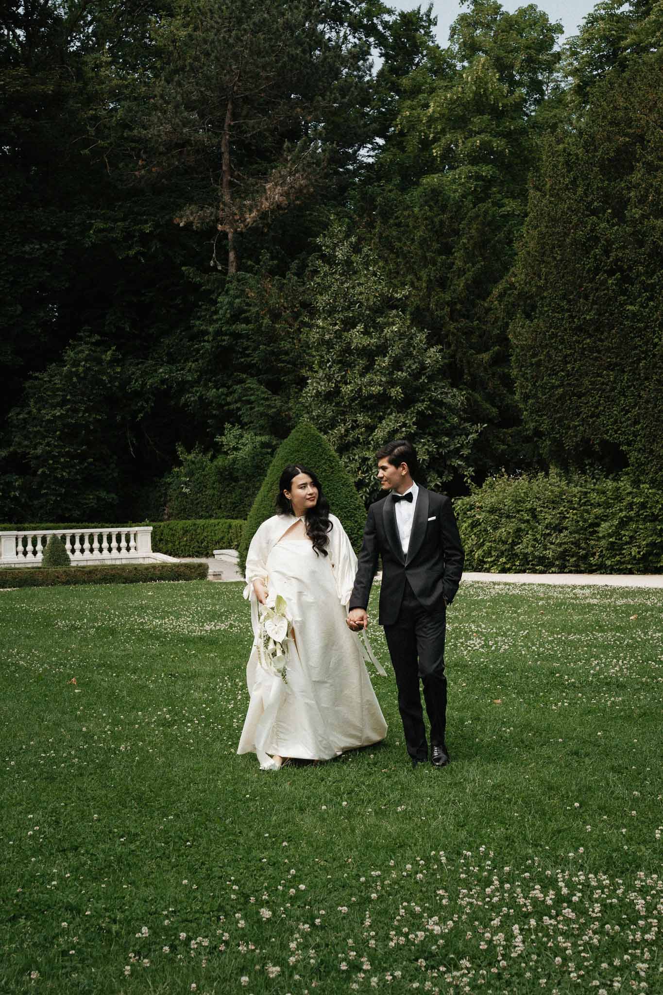 Bride in structured ball gown with cape and groom in black tuxedo walking hand in hand in formal chateau garden