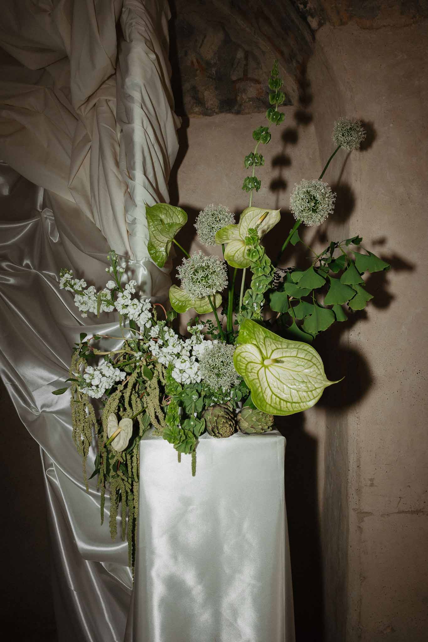 Sculptural green and white arrangement with anthurium, allium, and artichokes on white pedestal in stone chapel