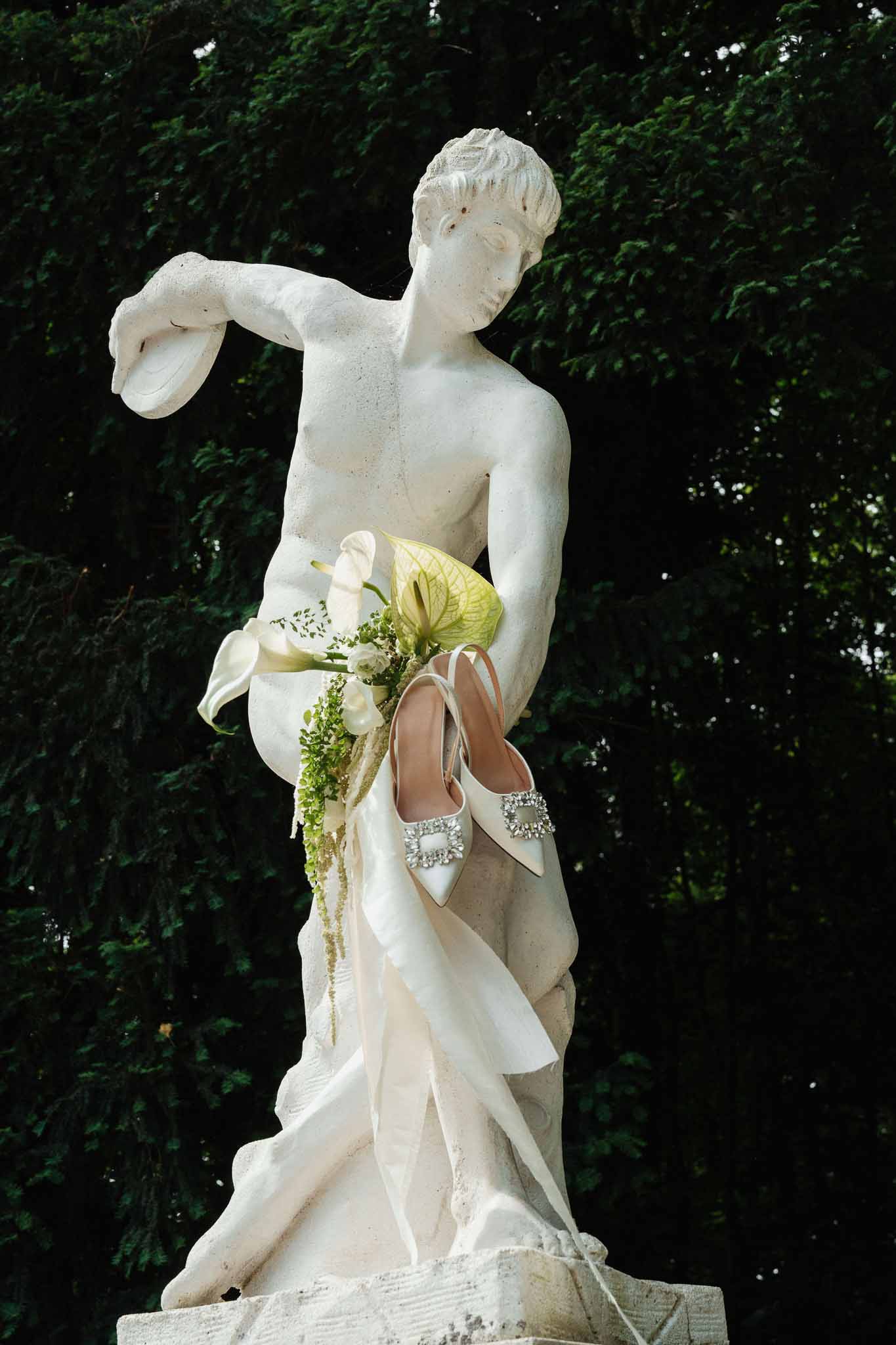 Ivory satin heels and bridal bouquet of white calla lilies with trailing amaranthus on classical stone statue
