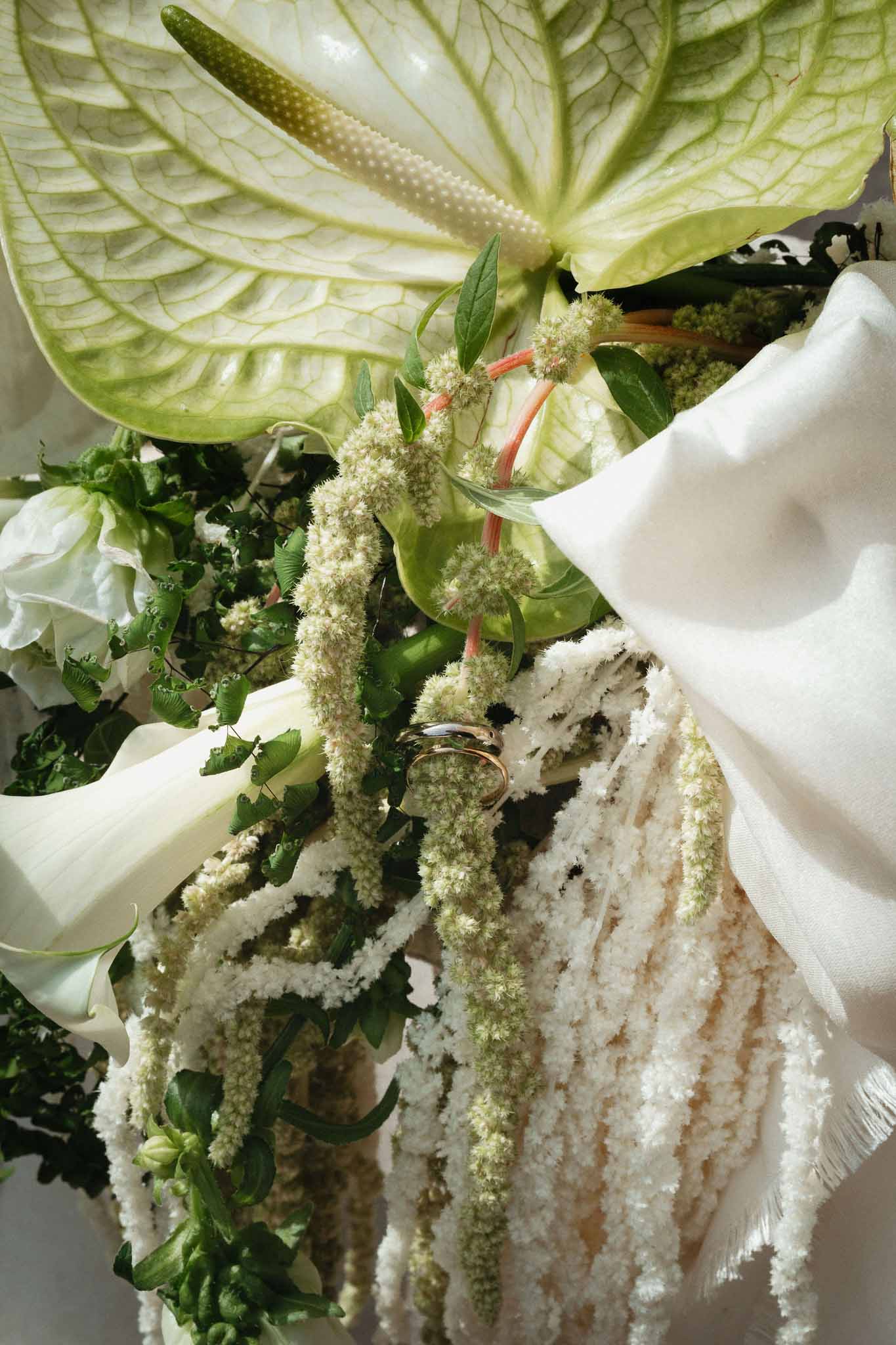 Modern bouquet of green-white anthurium, calla lilies, and trailing amaranthus with two gold wedding bands