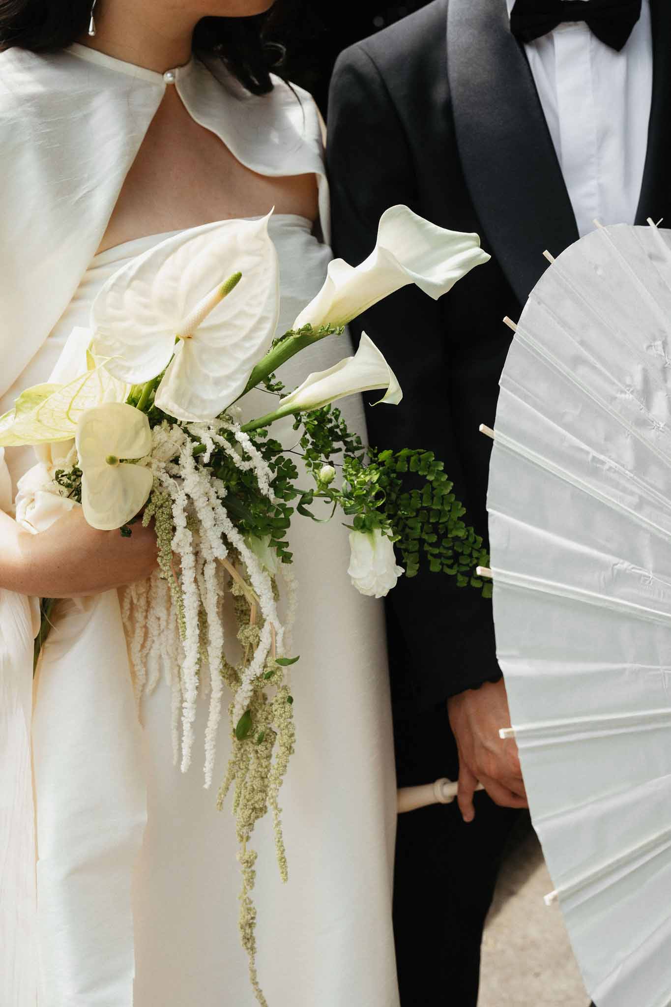 Close-up of bride holding cascading white anthurium and calla lily bouquet beside groom in black tuxedo