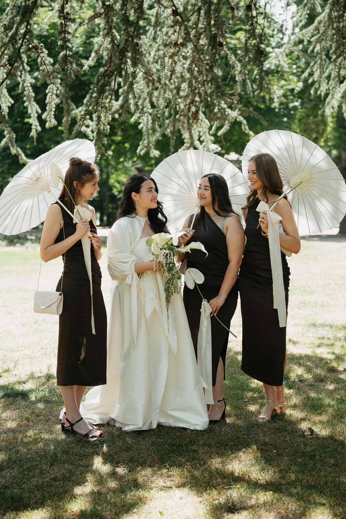 Bride in white gown with three bridesmaids in black midi dresses holding white paper parasols in garden