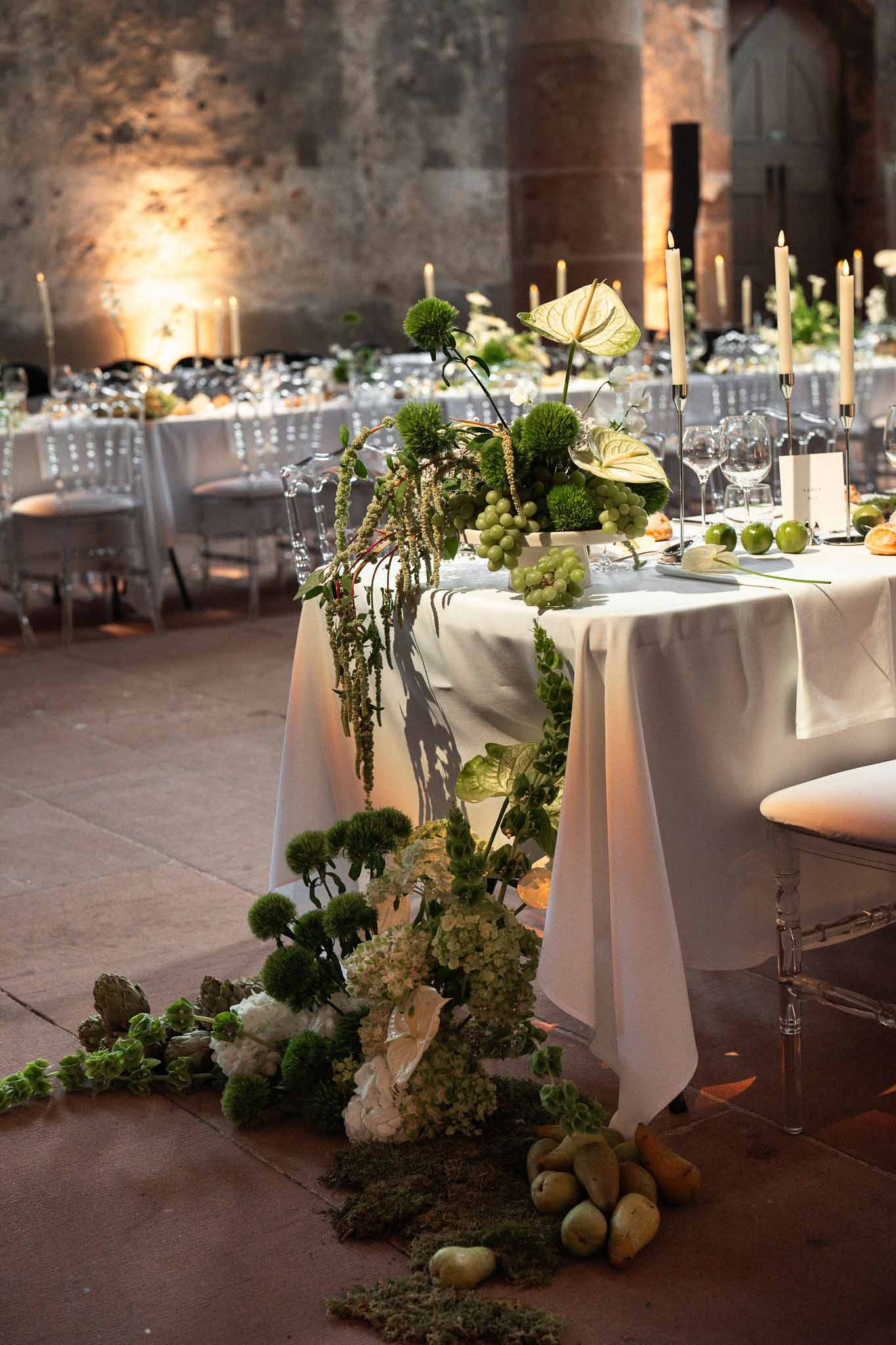 Botanical centerpiece of green anthurium, grapes, and artichokes with gold tapers in stone-walled venue