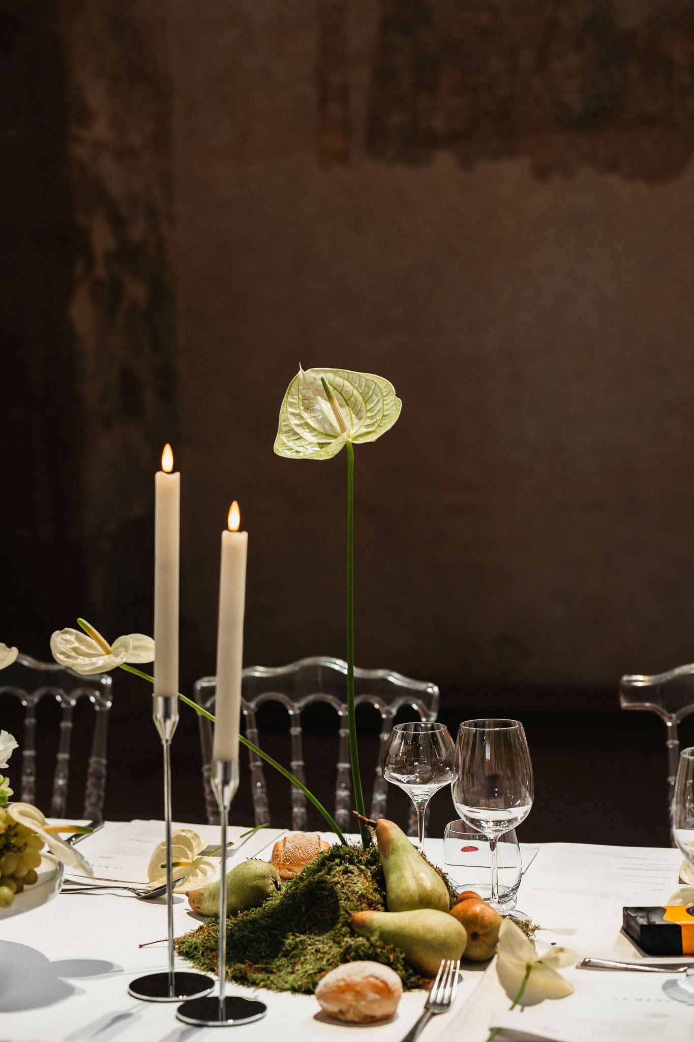 Modern botanical centerpiece with green pears, moss, and anthurium beside ivory taper candles on white linen