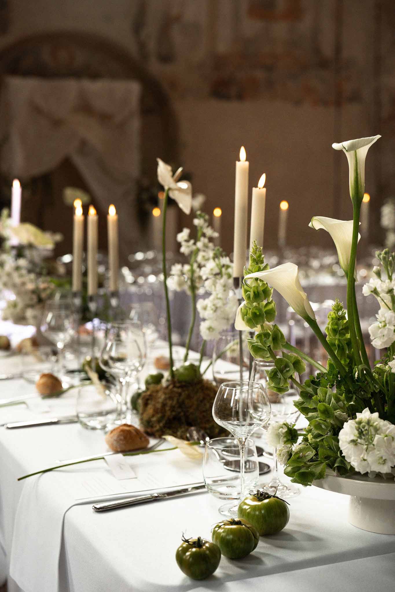 Reception tablescape with white calla lilies, green blooms, tall taper candles, and heirloom tomatoes in stone venue
