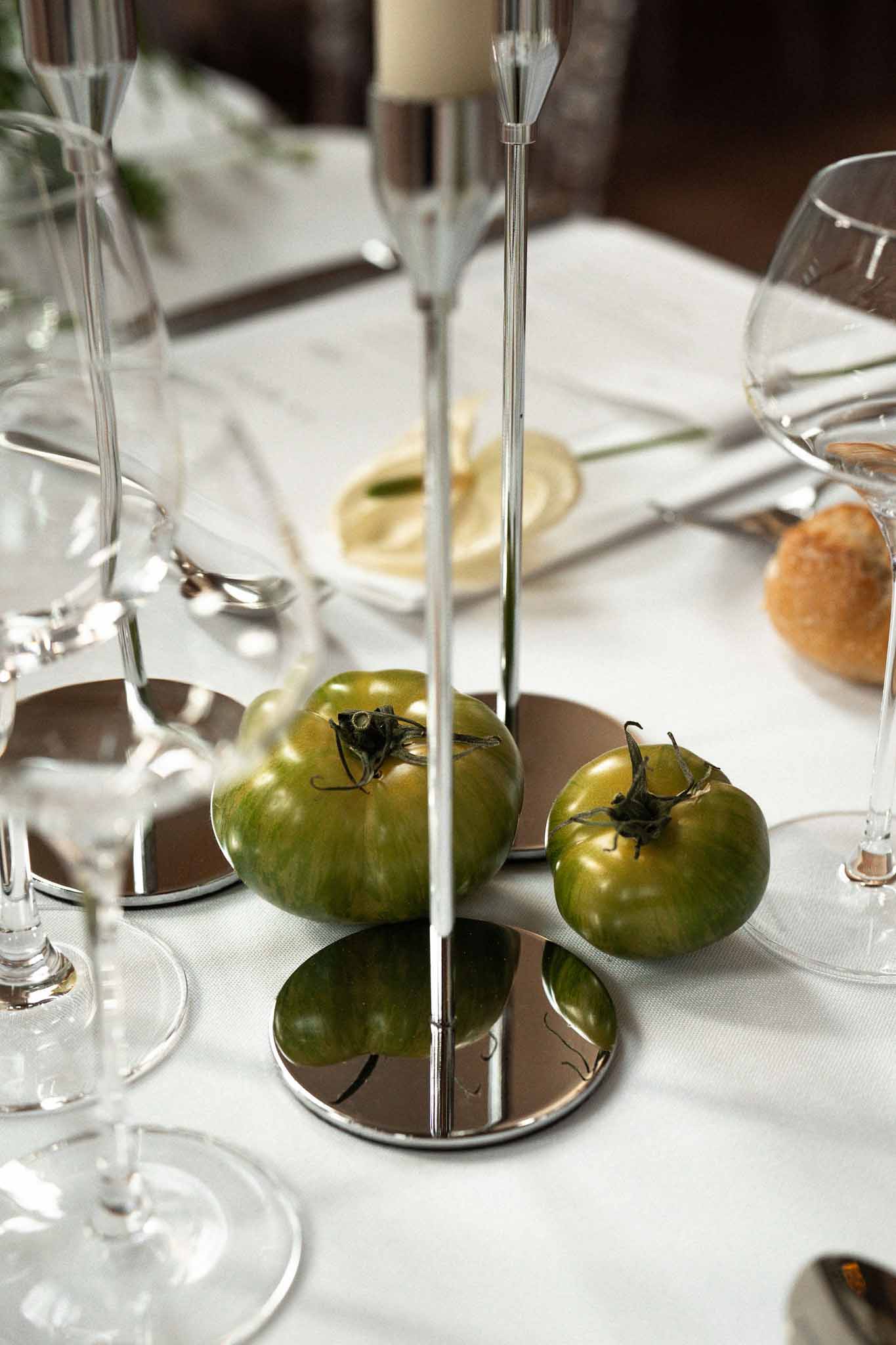Three green heirloom tomatoes around chrome candlestick with cream taper on mirrored base at table setting