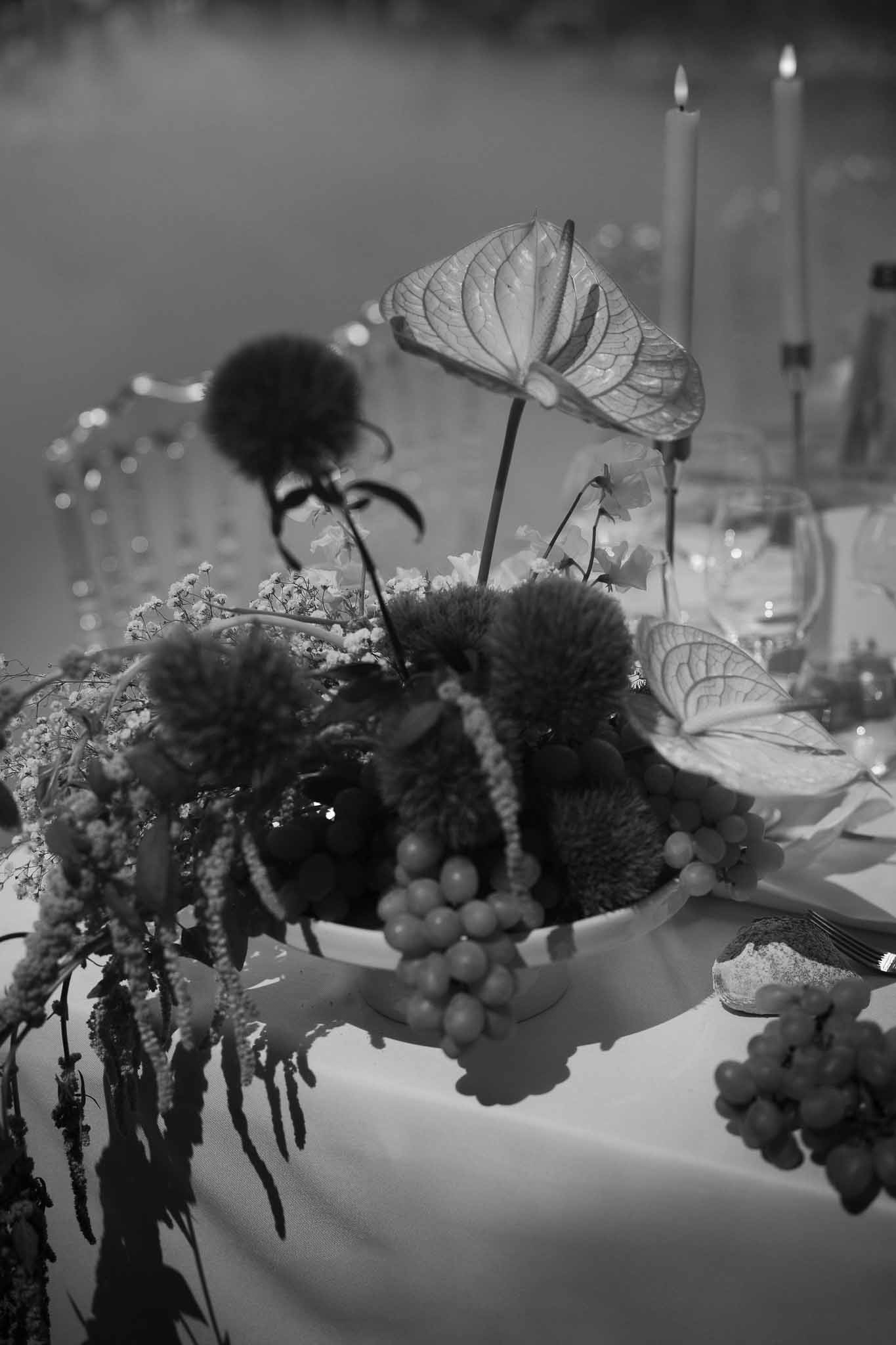 Black and white close-up of reception centerpiece with anthurium, dianthus, amaranthus and grapes in pedestal bowl