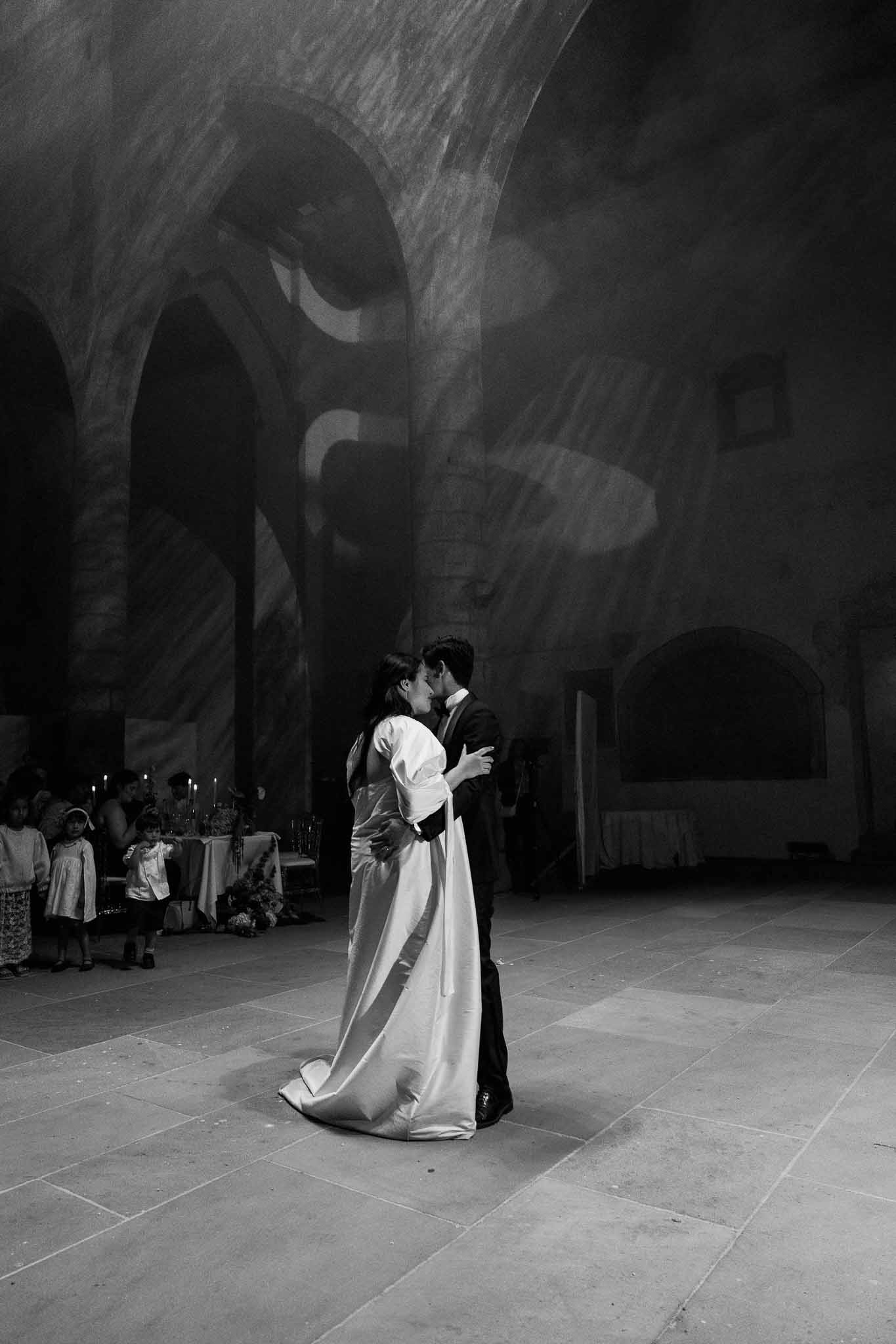 Black and white first dance kiss in stone-vaulted hall with ribbed arches and light projections