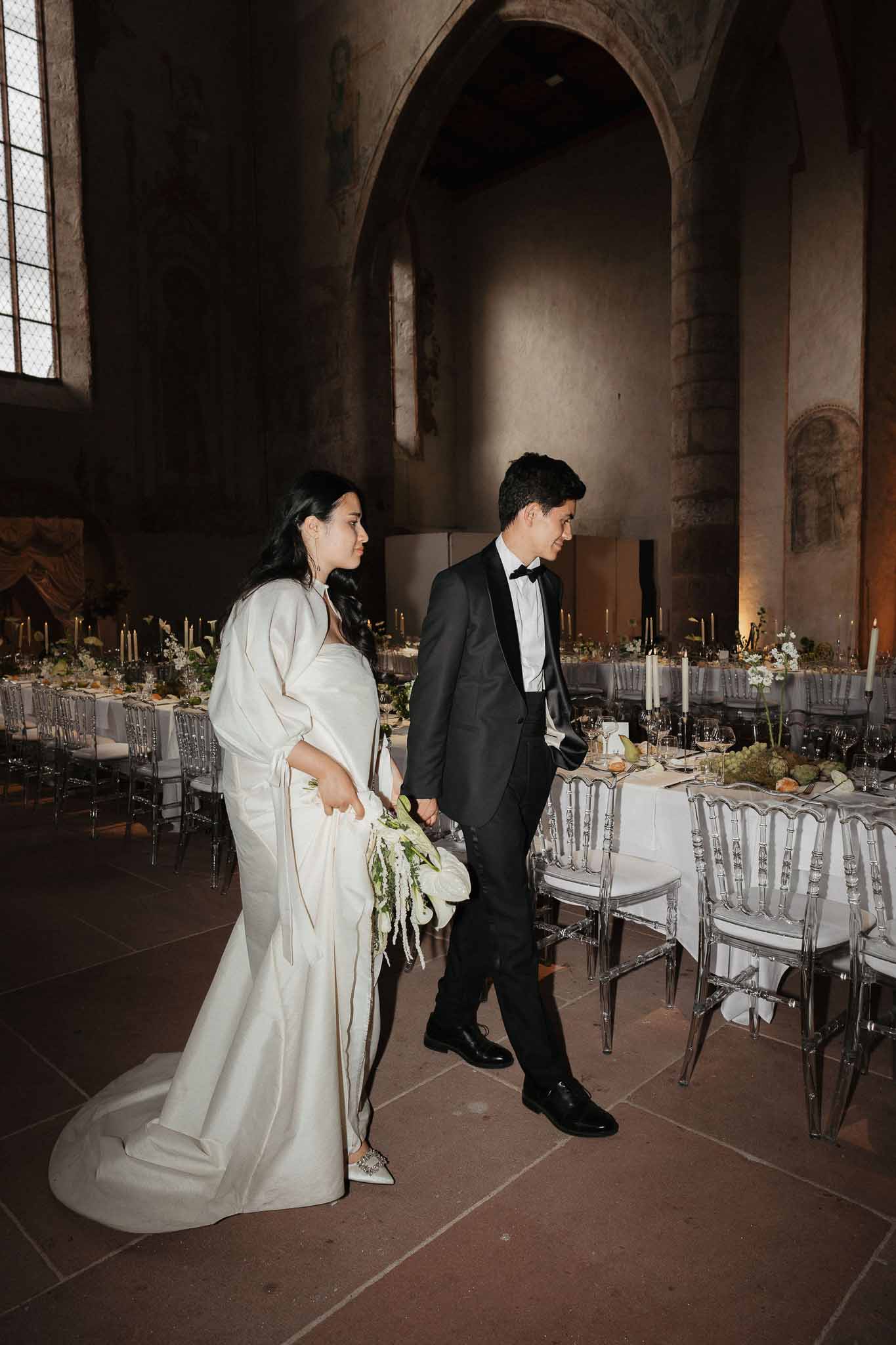 Bride and groom walk through medieval abbey reception hall with white banquet tables, ghost chairs, and taper candles