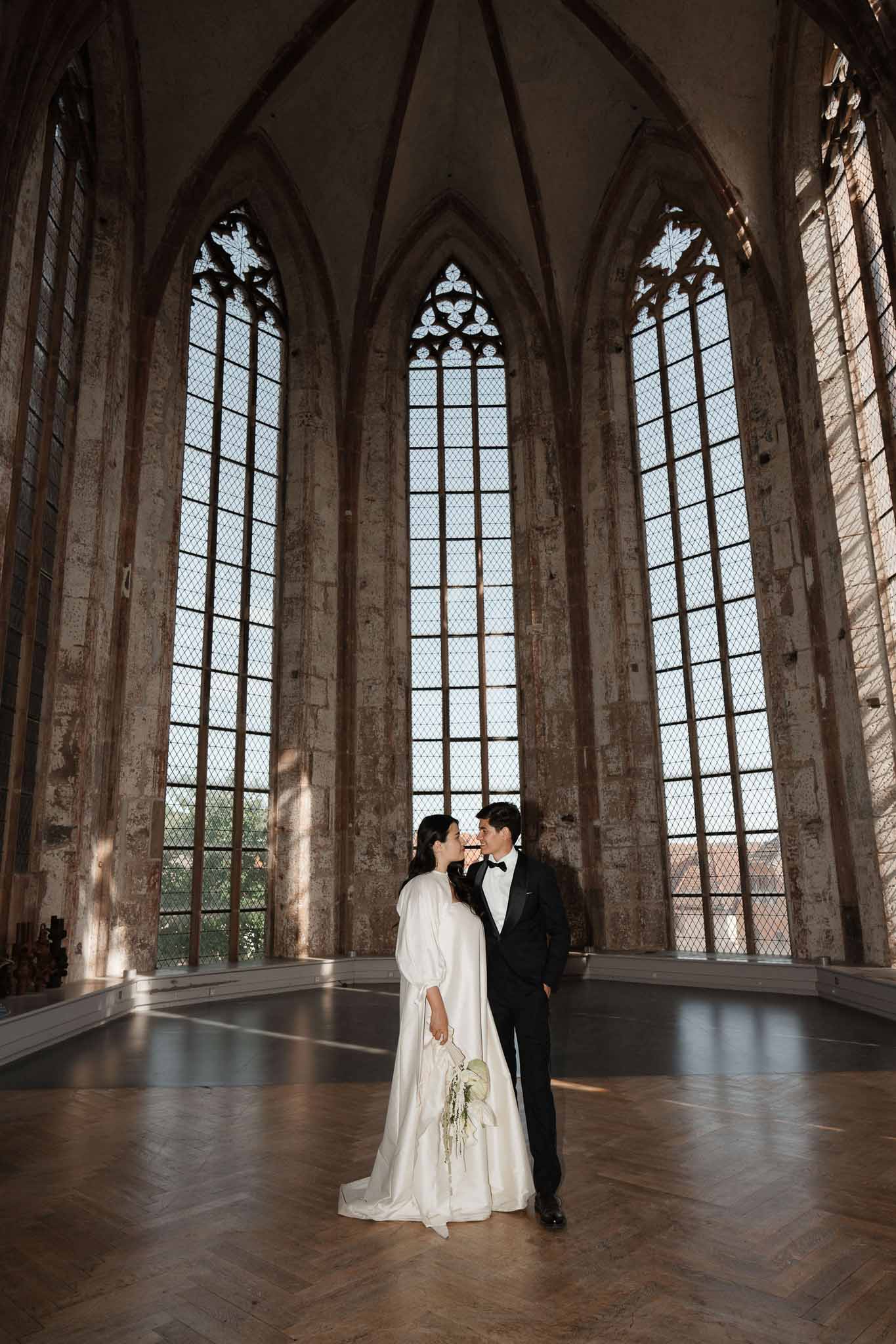 Bride and groom touching foreheads inside Gothic chapel with tall lancet windows and parquet floor