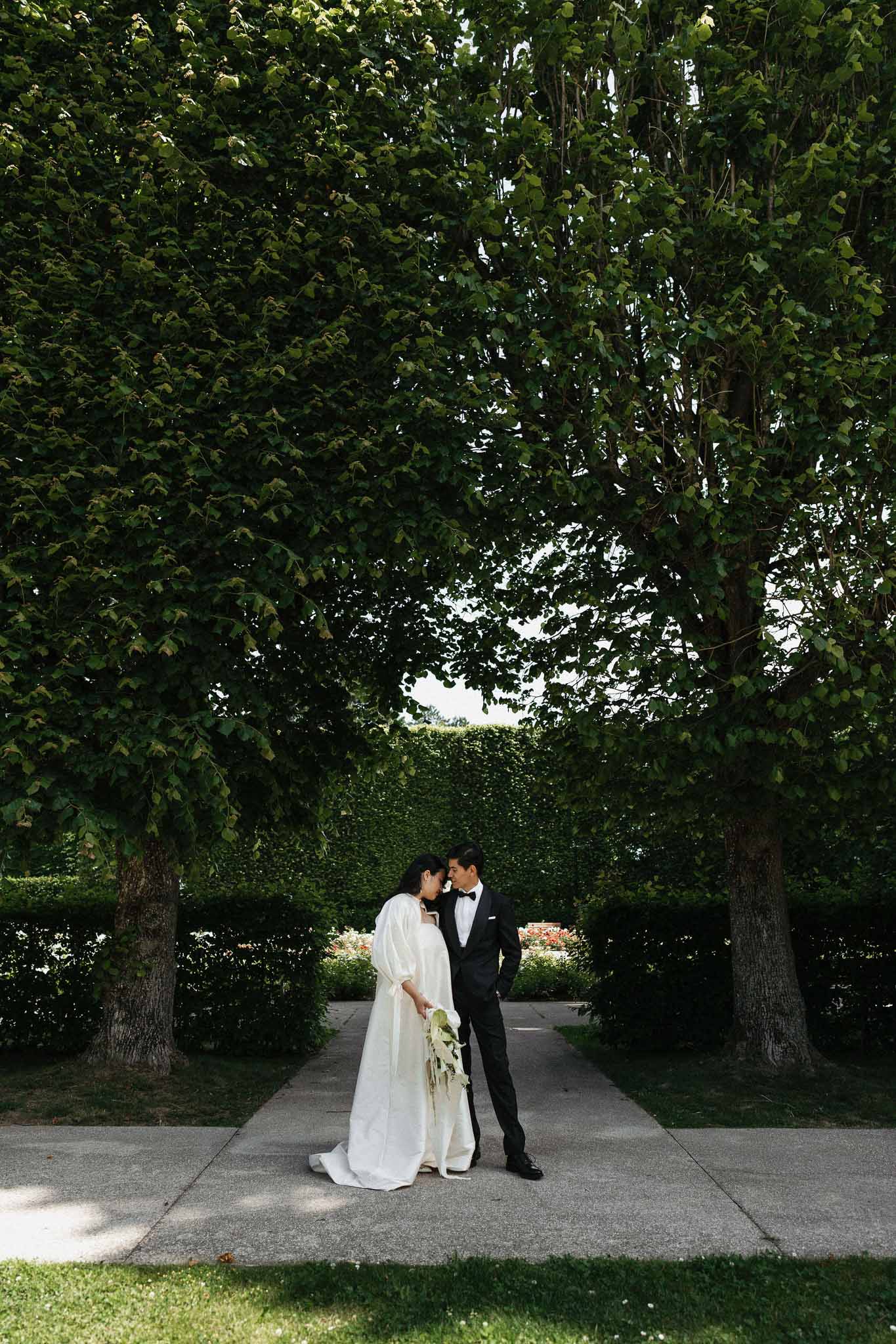 Bride in puff-sleeve ivory gown and groom in tuxedo face-to-face between tall hedged garden trees