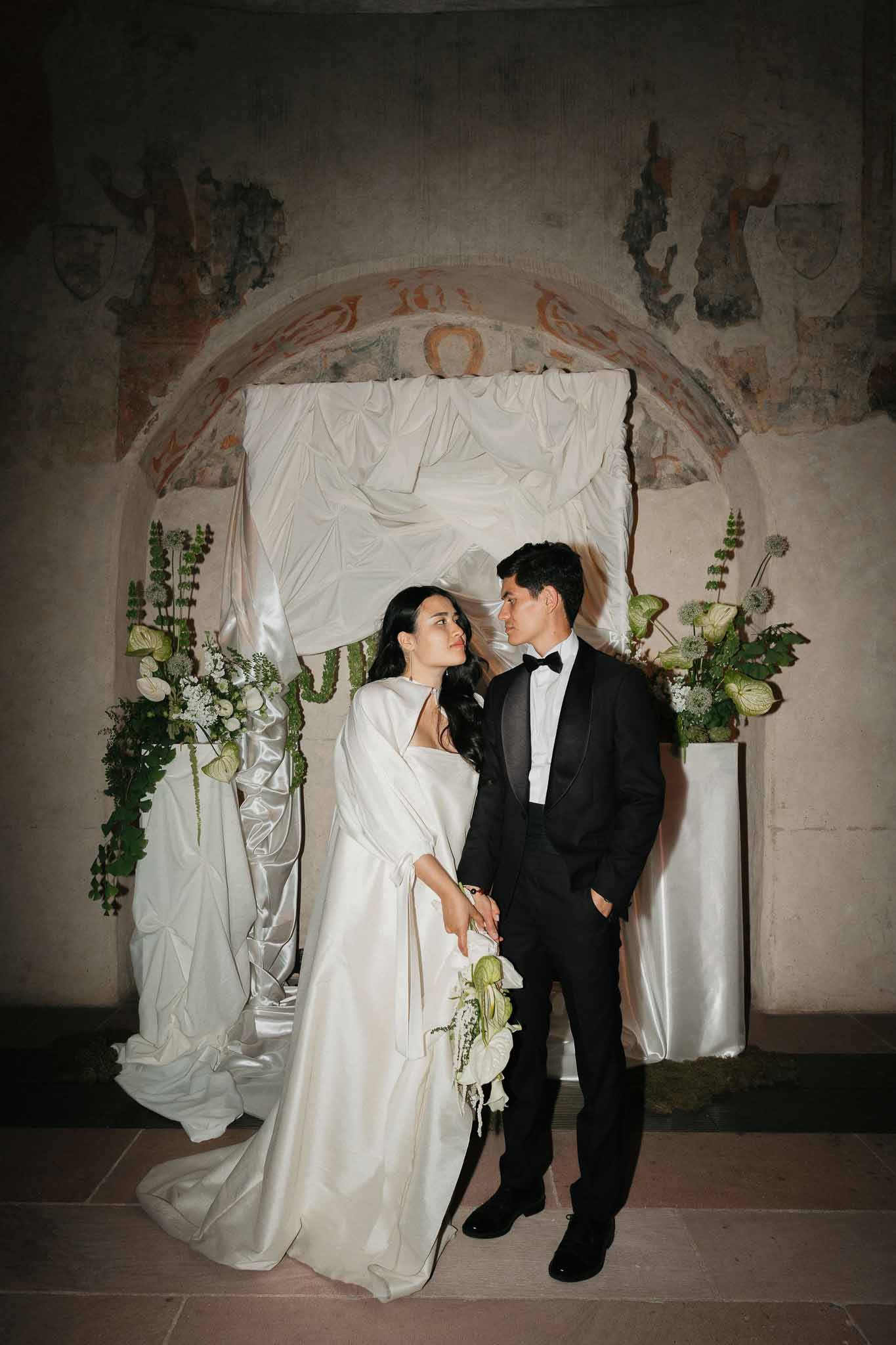 Bride and groom facing each other in historic chapel with white anthurium arrangements on pedestals
