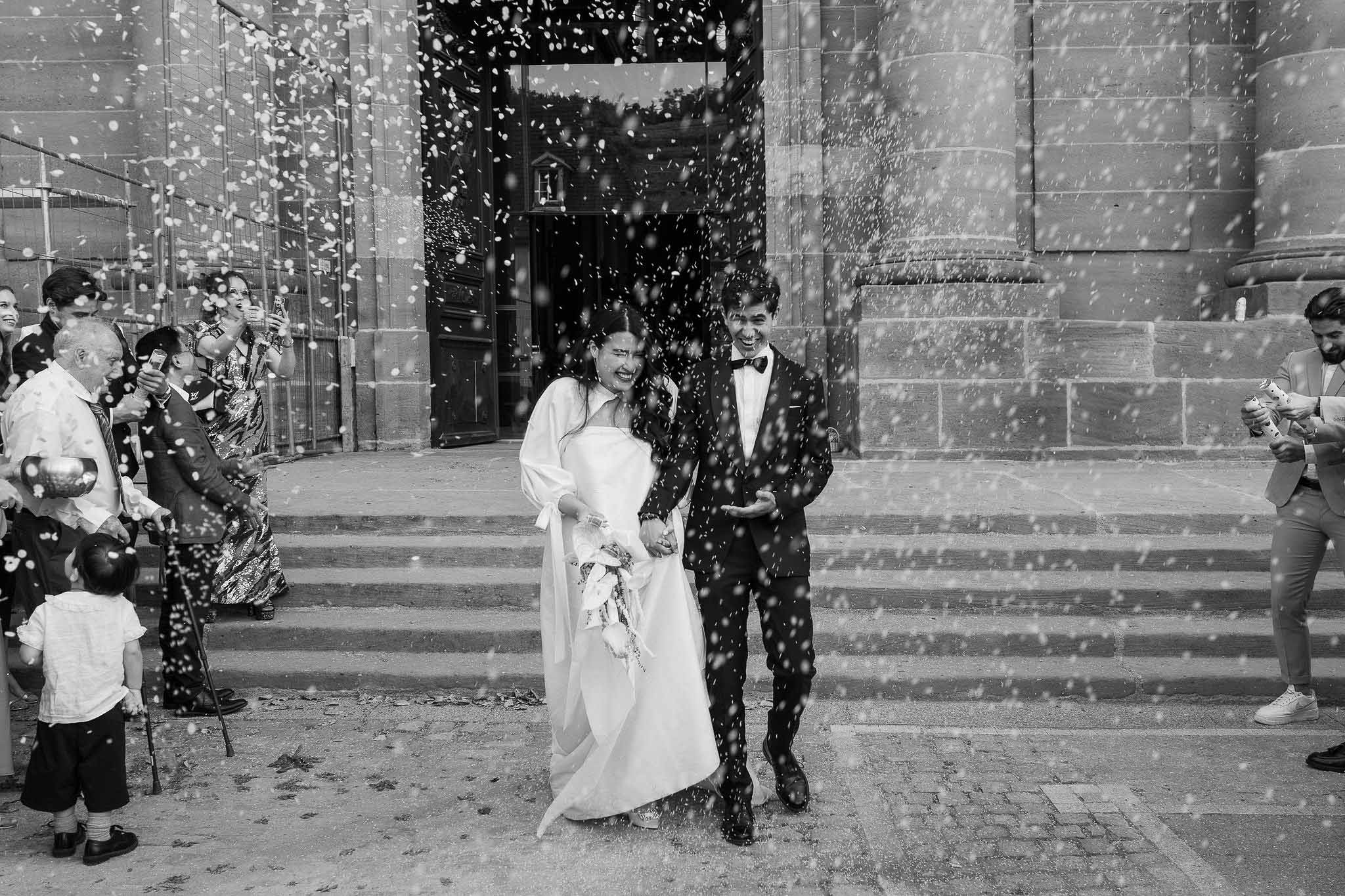 Black-and-white shot of bride and groom exiting stone church to a shower of rice with laughing guests on steps