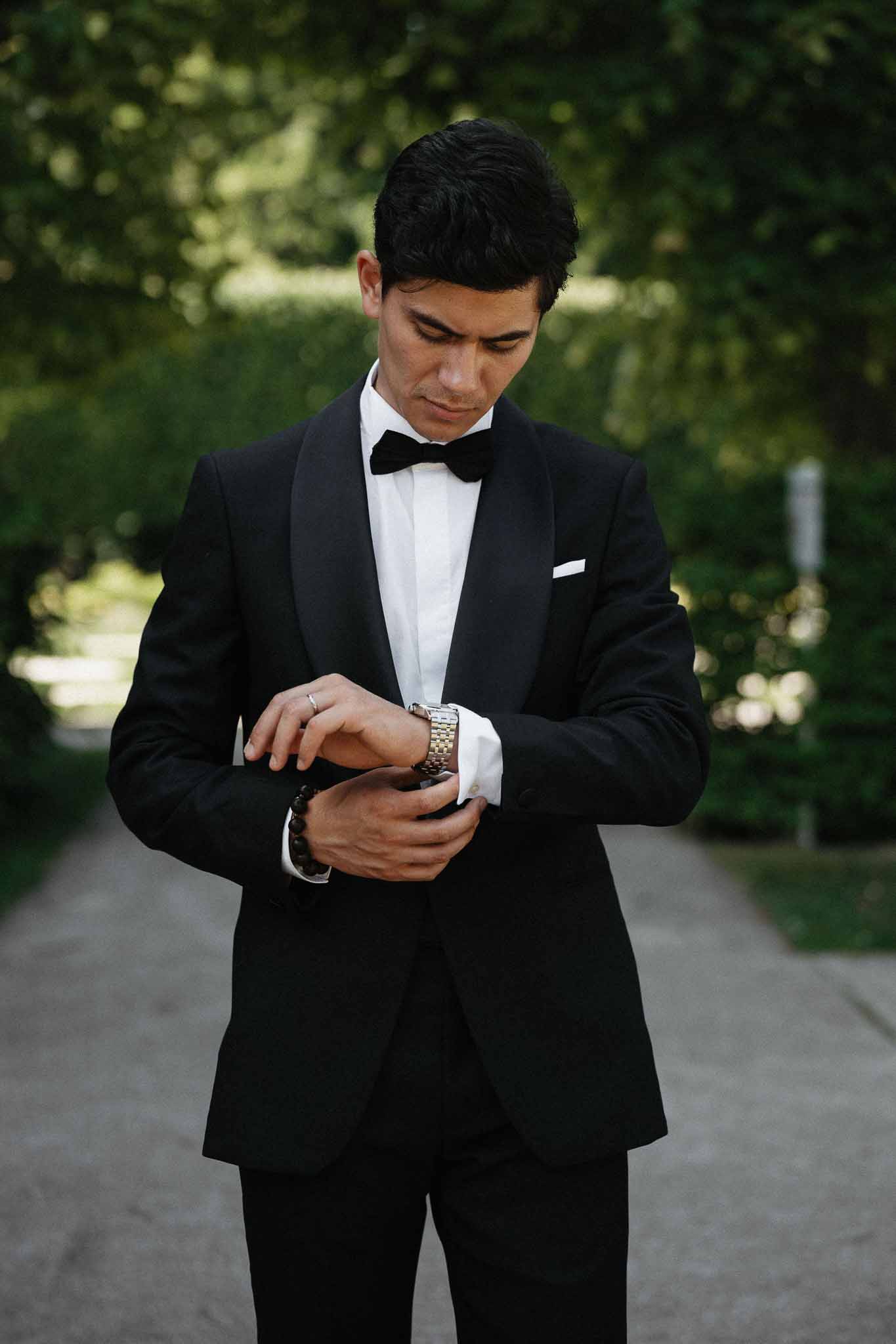 Groom in black shawl-lapel tuxedo adjusting cufflinks on tree-lined garden pathway