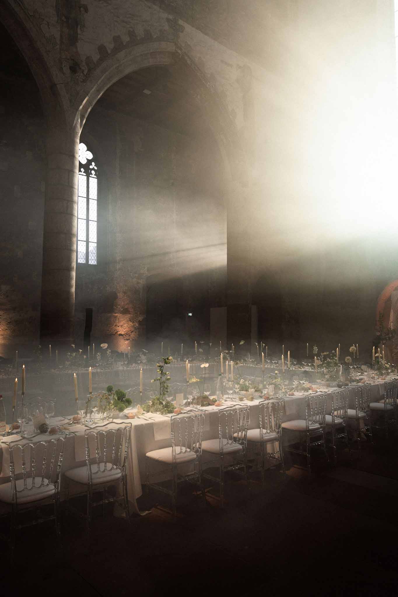 Medieval stone abbey reception with two banquet tables, taper candles, and dramatic light through gothic window