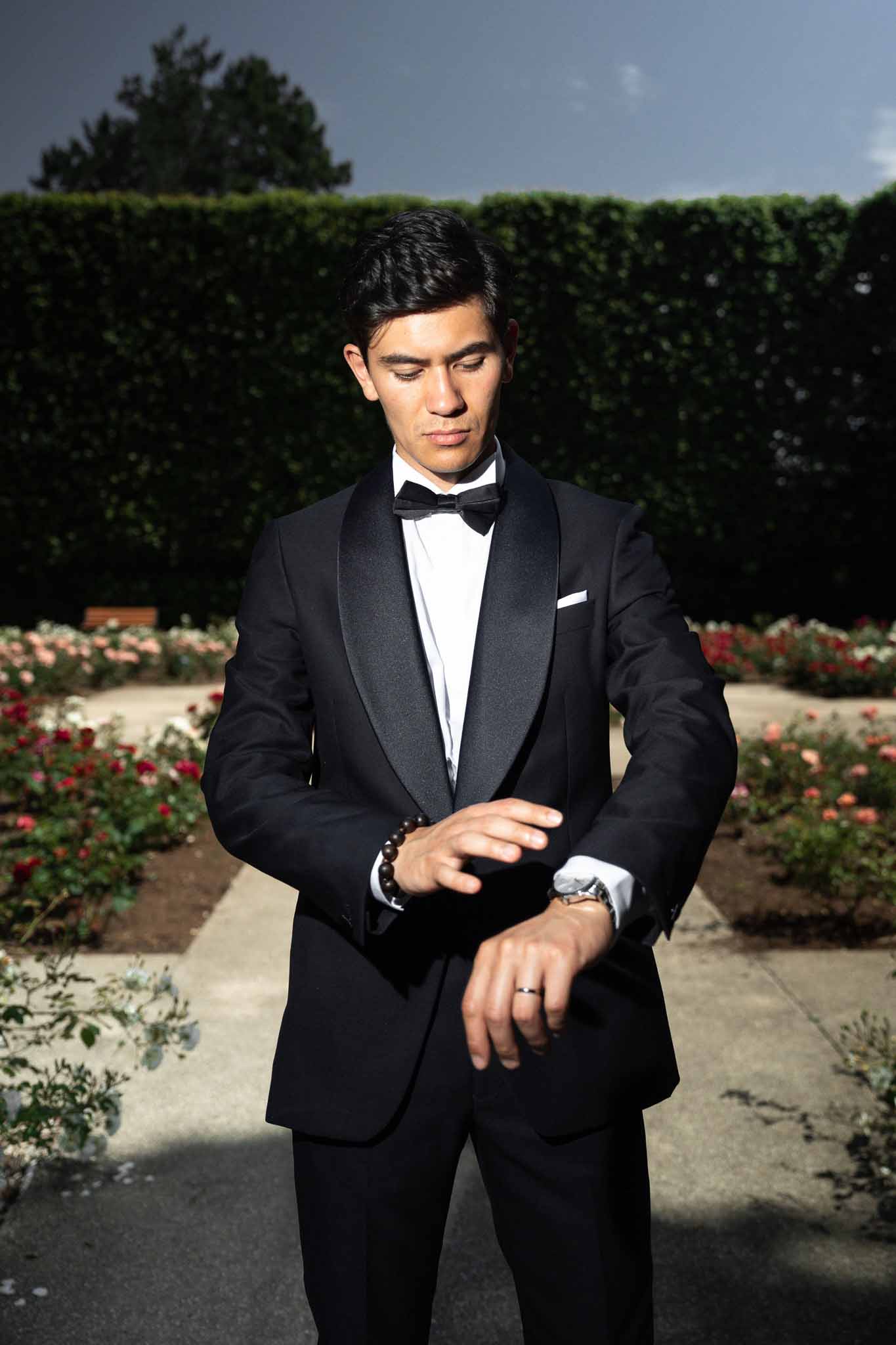 Groom in black tuxedo with bow tie adjusting cufflinks in a formal garden with rose beds