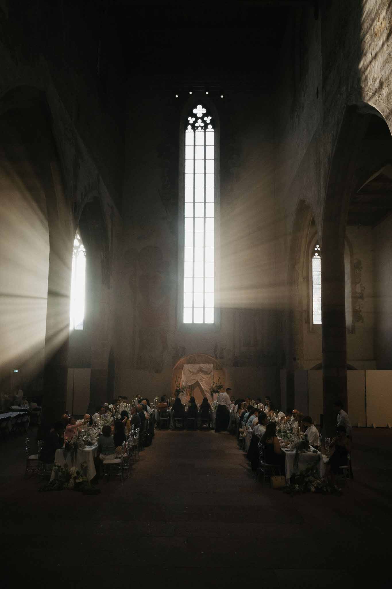 Wedding reception with long banquet tables and seated guests inside Gothic chapel with light beams