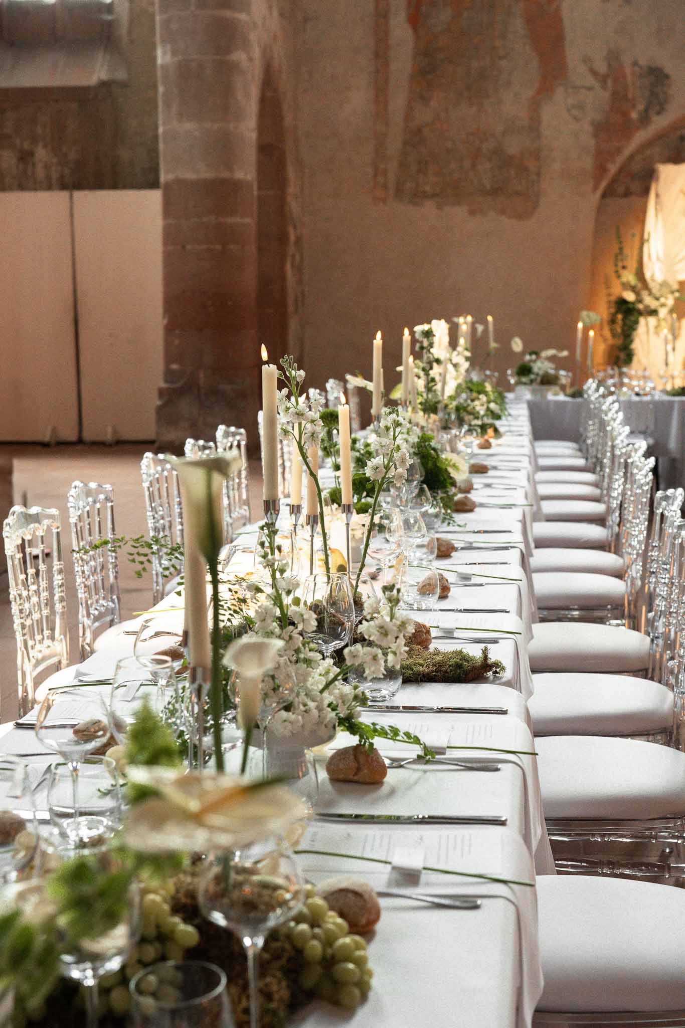 Long reception table with white calla lilies, moss, grapes, and taper candles inside a stone-walled venue with frescoes
