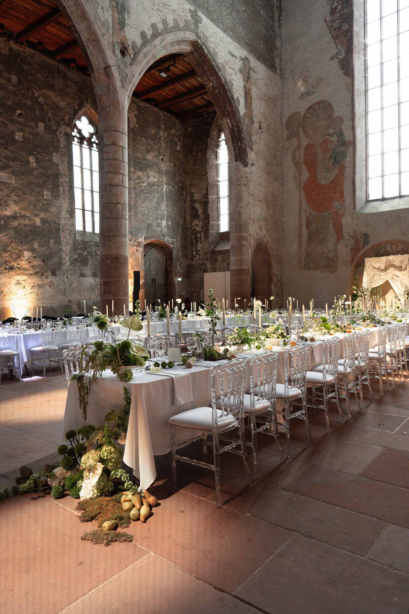 Medieval stone chapel reception with long tables, ghost chairs, green botanical runners, and taper candles