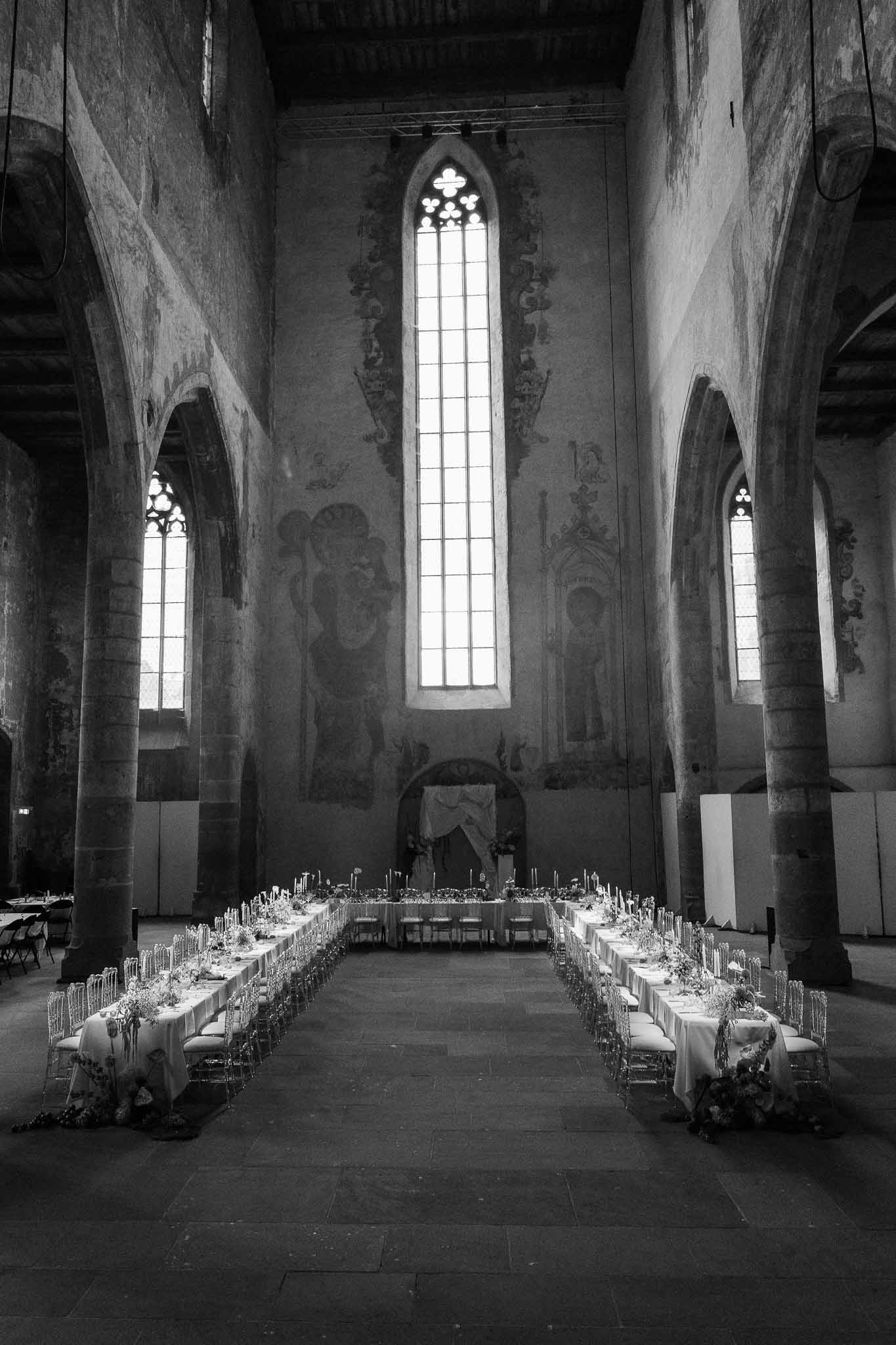 Black-and-white reception setup inside Gothic abbey nave with long banquet tables, candles, and vaulted stone ceiling