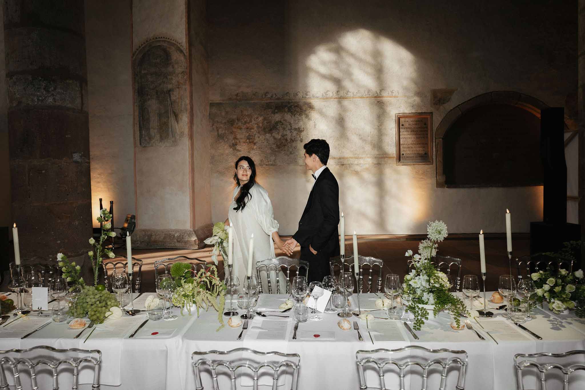 Bride and groom standing behind long banquet table in ancient chapel with white floral garland and taper candles