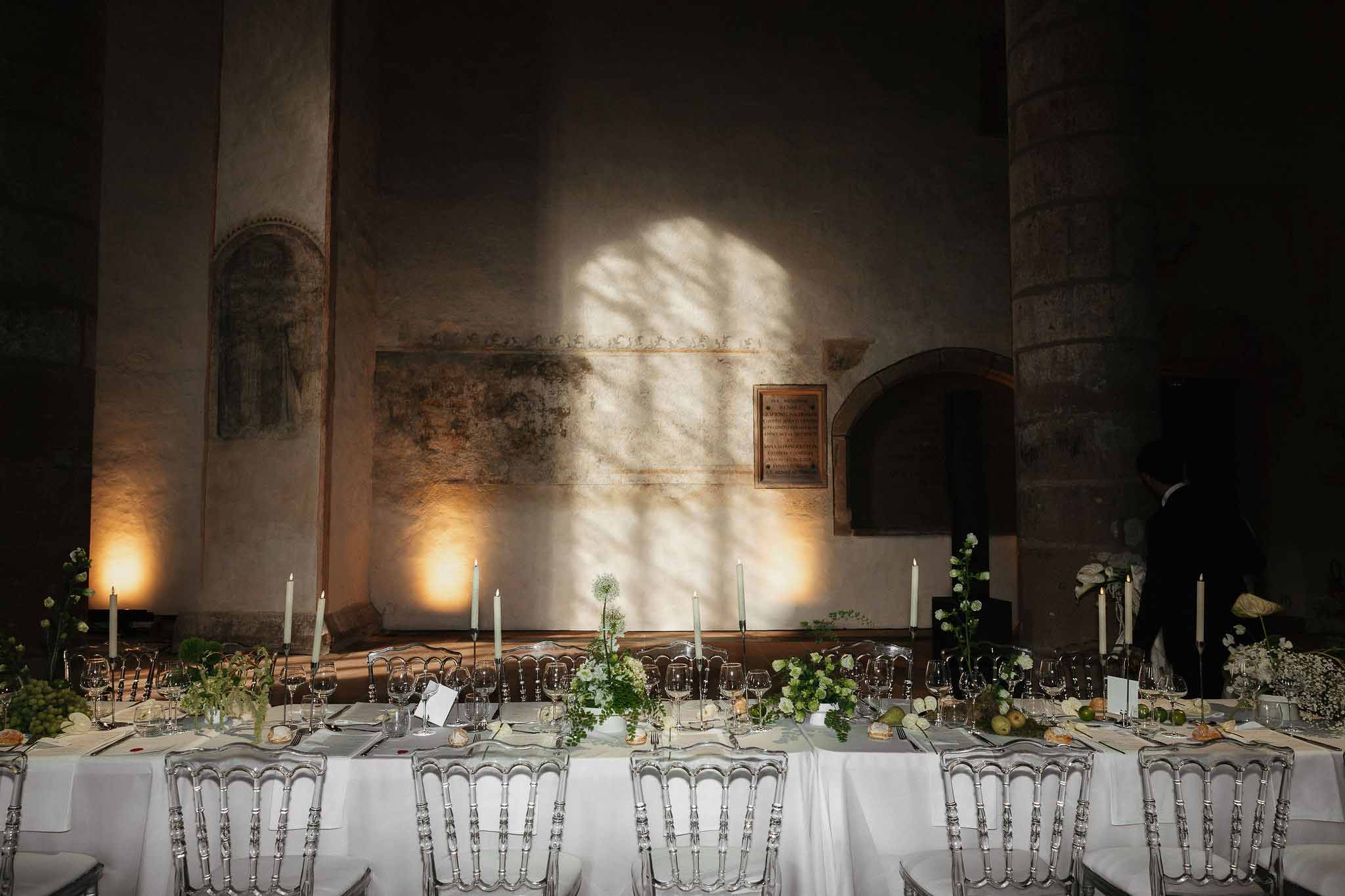 Long banquet table in historic chapel with white florals, taper candles, acrylic chairs, and fresco walls