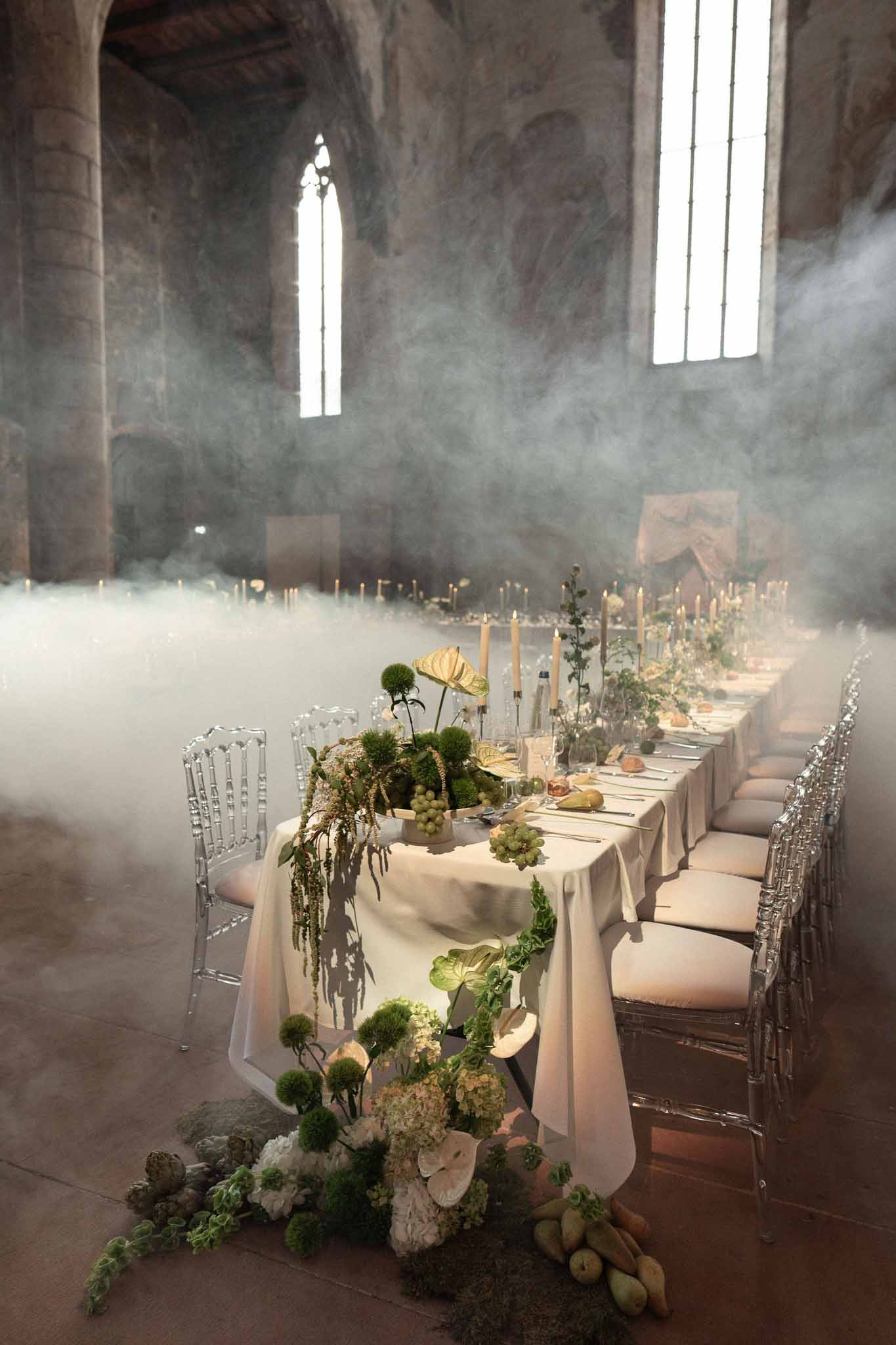 Reception tablescape in Gothic stone hall with dry-ice mist, green floral centerpieces, ghost chairs, and taper candles