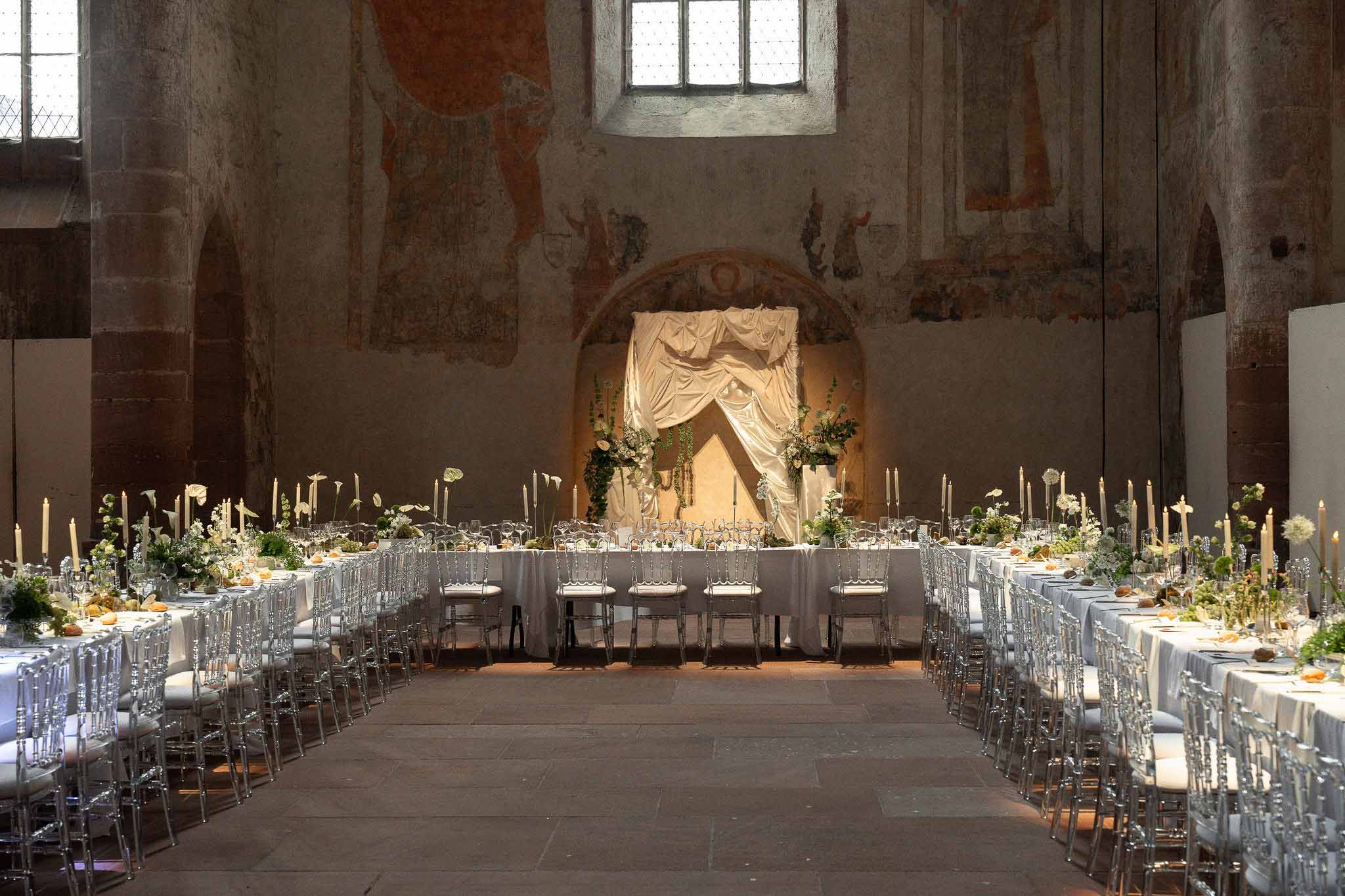 Wedding reception setup inside medieval chapel with long banquet tables, ghost chairs, calla lilies, and draped head table