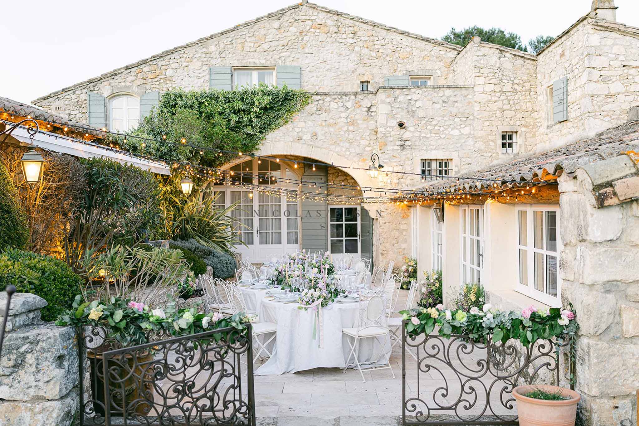 Dusk courtyard reception with round tables, blush centerpieces, and wrought-iron gates with garlands