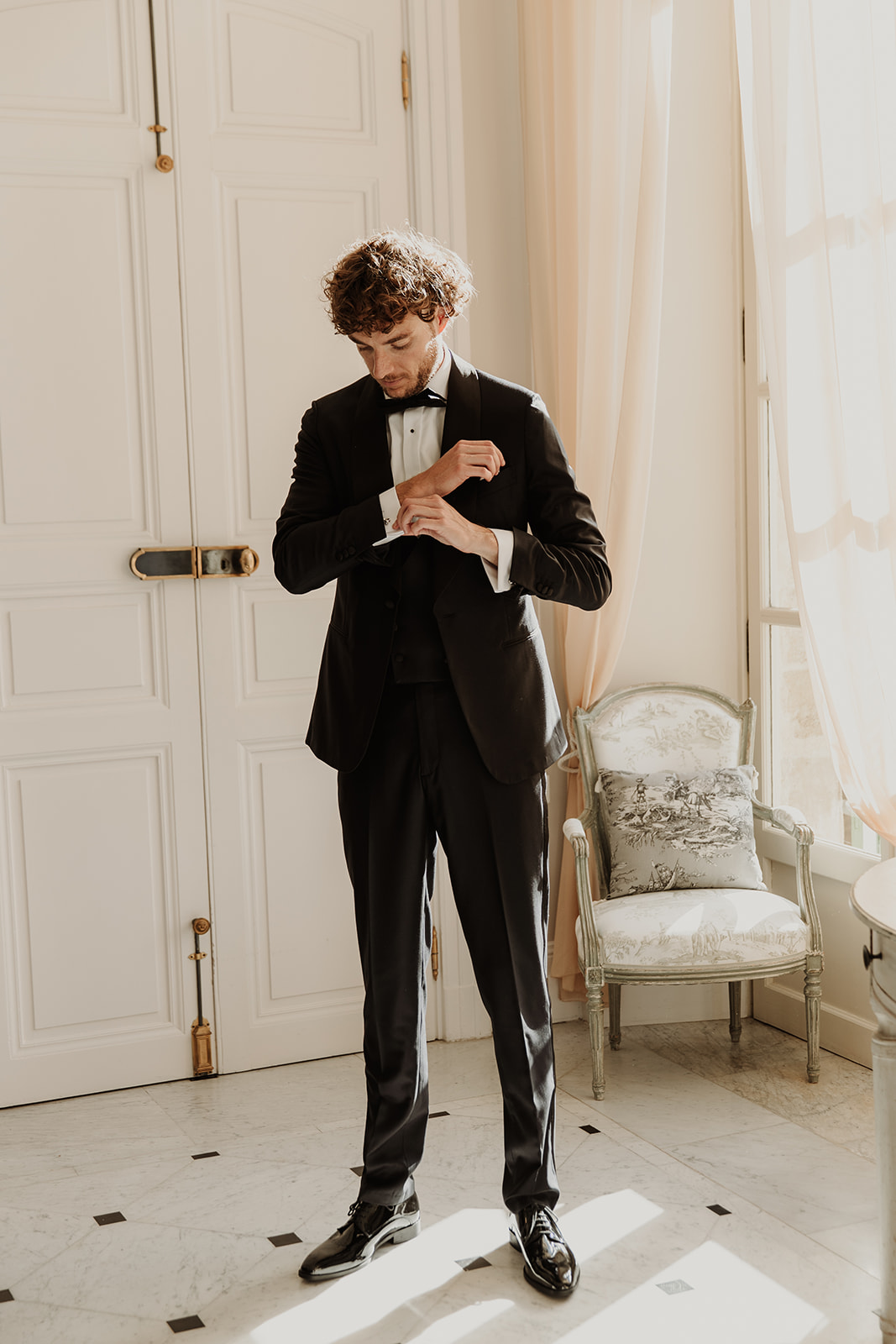 Groom adjusting cufflinks in black tuxedo inside ornate chateau room with marble floor and period furniture
