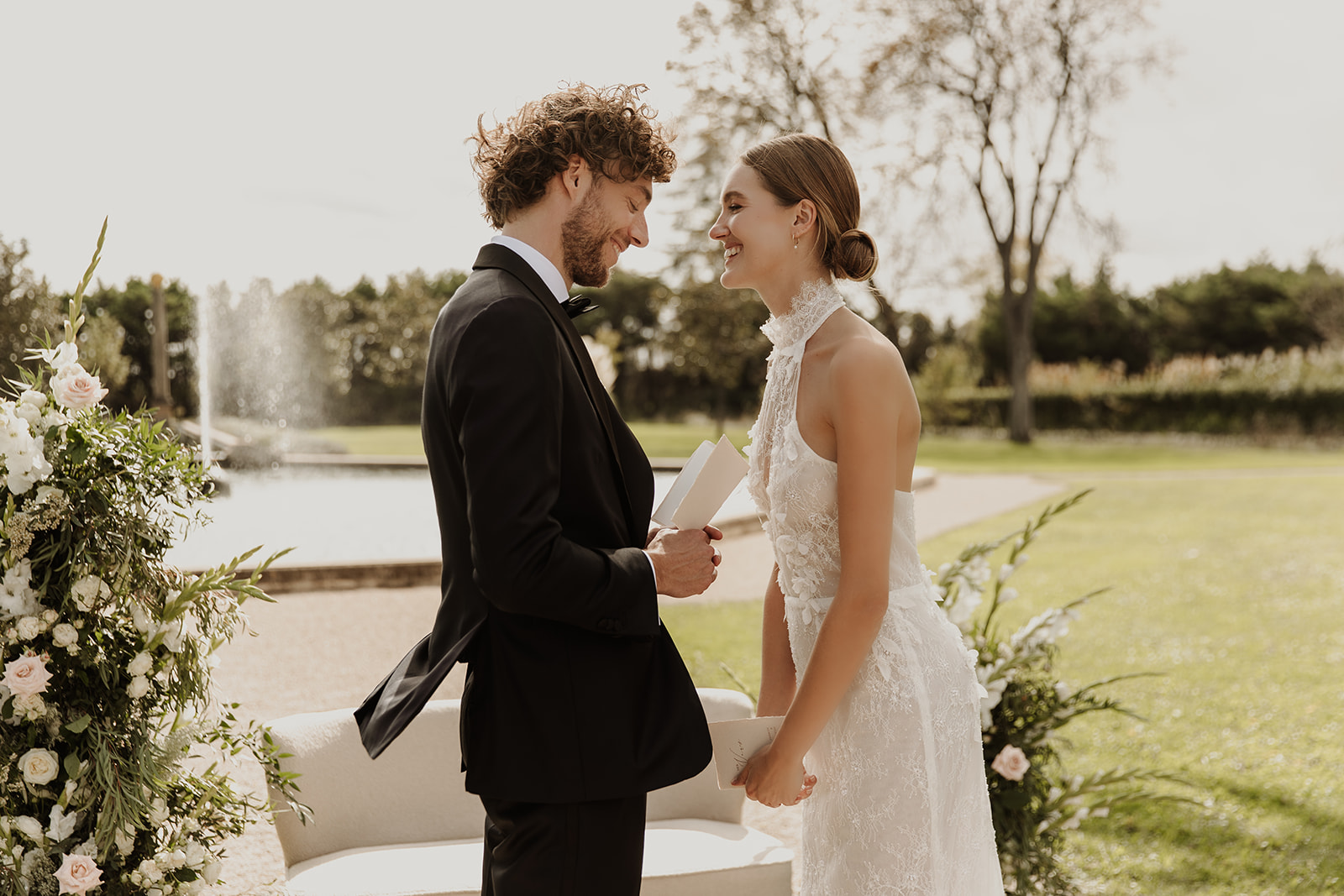 Groom reads vows holding bride's hands beside white and blush rose arrangements in garden setting