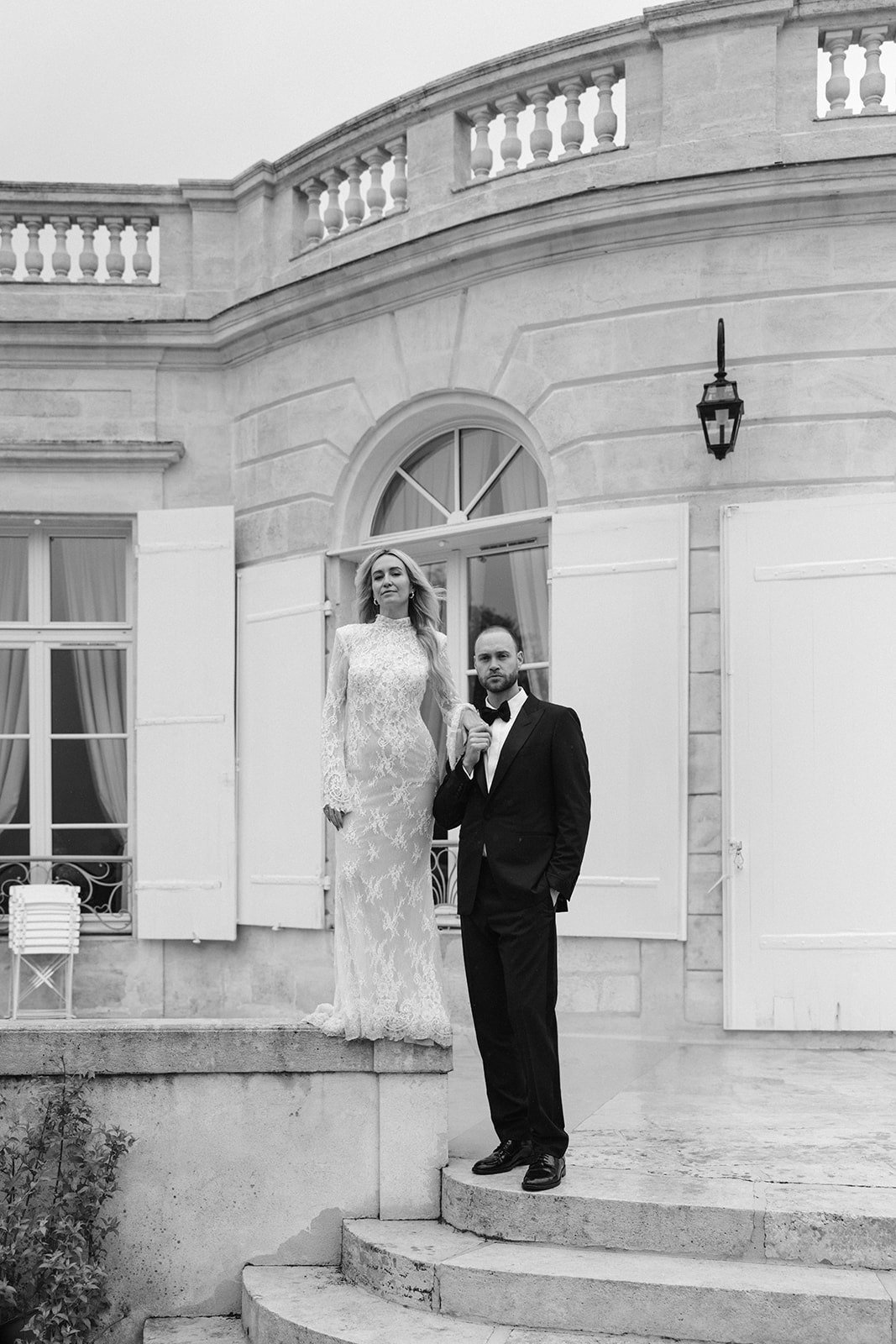 Black and white couple on chateau steps, bride in allover lace gown, groom in tuxedo facing camera