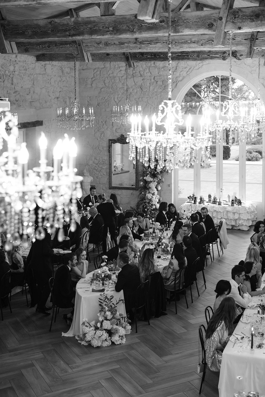 Black and white wide shot of formal reception dinner in a chateau with crystal chandeliers and long banquet tables